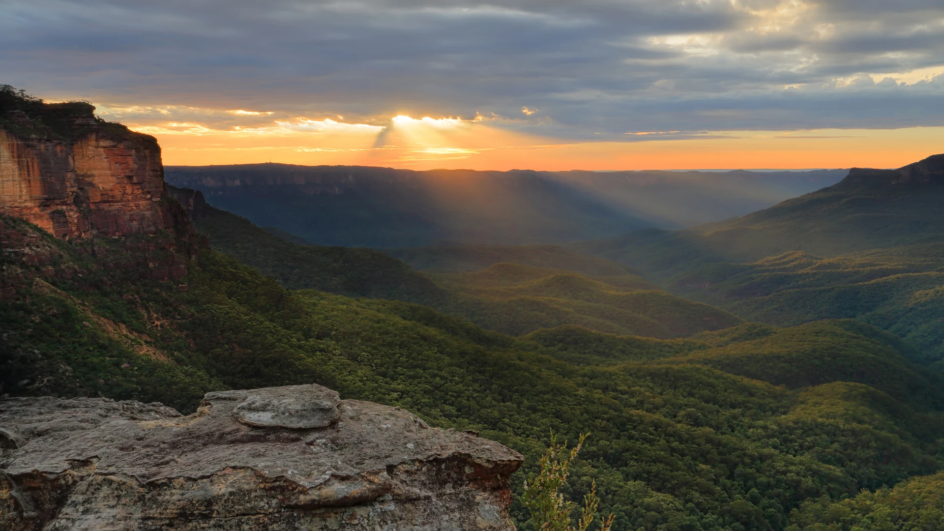 Sunset rays break through clouds over a lush green mountain valley with rocky cliffs in the foreground.