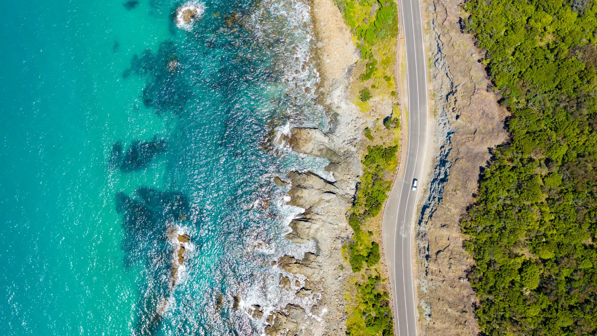 Aerial view of a coastal road winding along rocky shoreline with turquoise ocean on one side and green vegetation on the other.