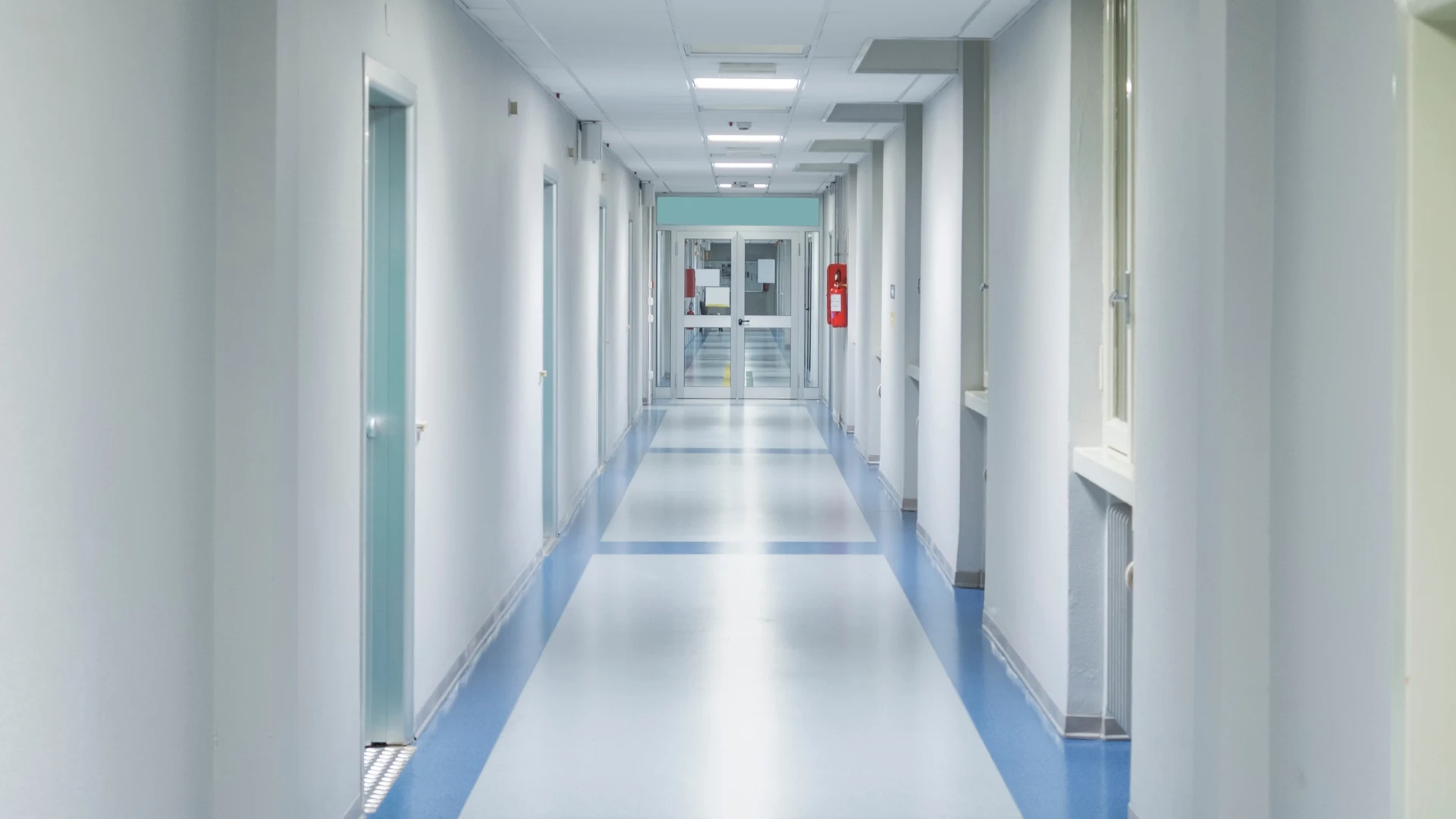 Empty hospital corridor with blue and white flooring, closed doors on the left, windows on the right, and glass double doors at the end.