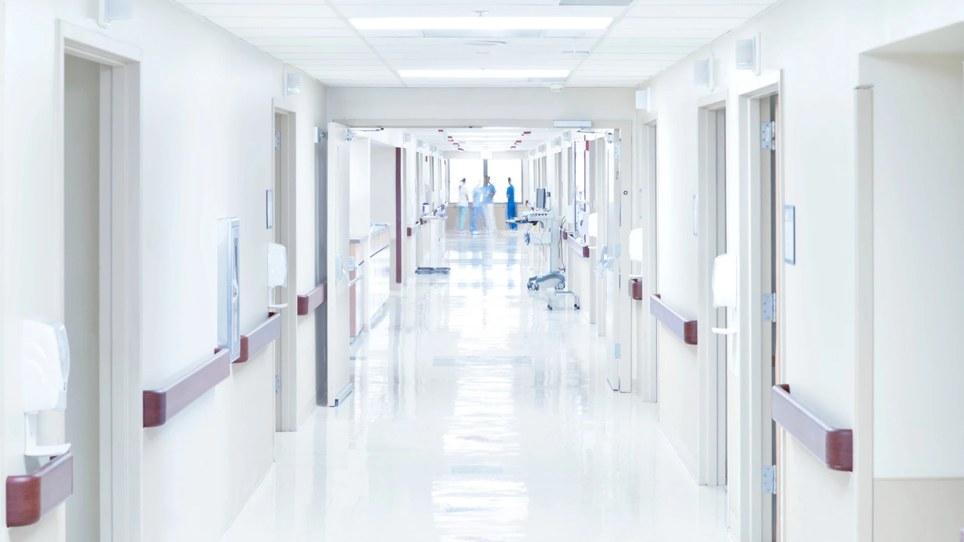 Bright hospital corridor with open doors and medical staff talking in the background.