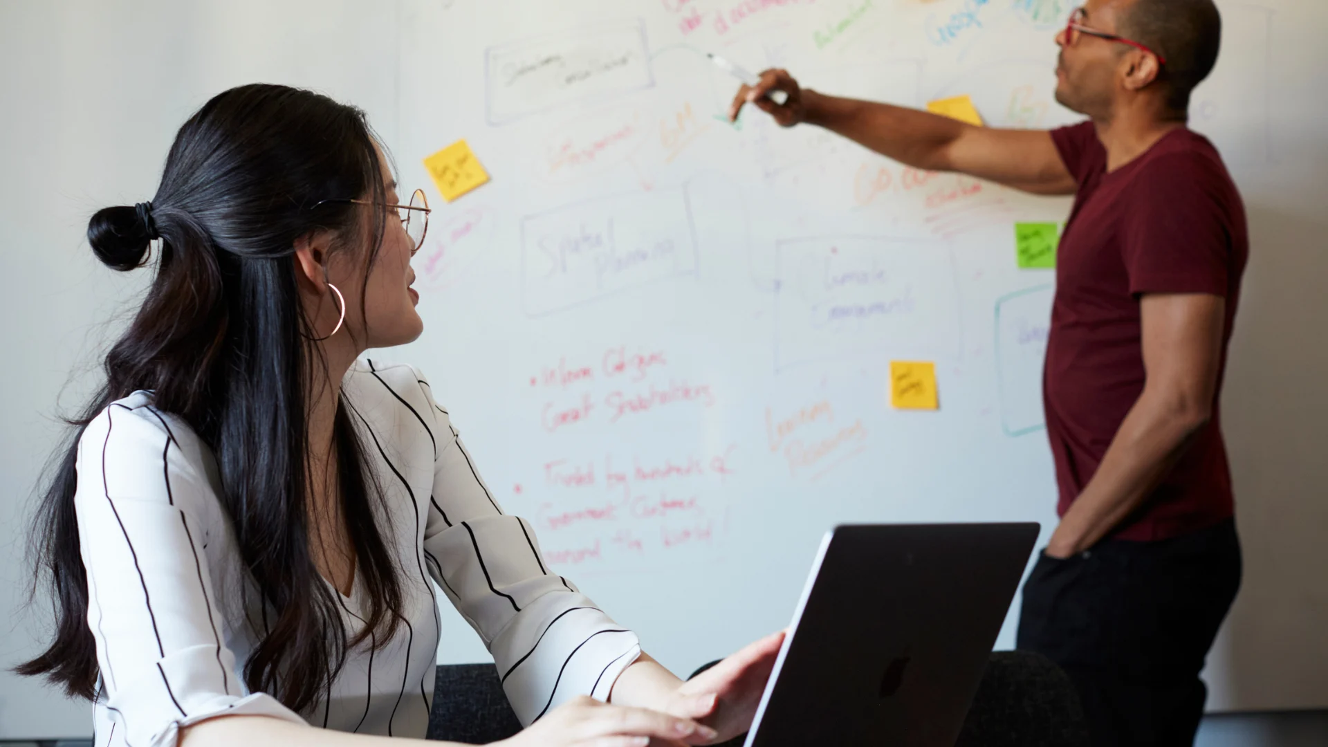 Man pointing at a whiteboard with notes while a woman with a laptop listens attentively in a meeting room.
