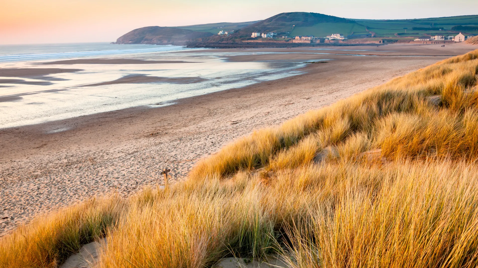 Golden dunes with tall grass overlooking a wide sandy beach and a coastal village under a clear sky at sunset.