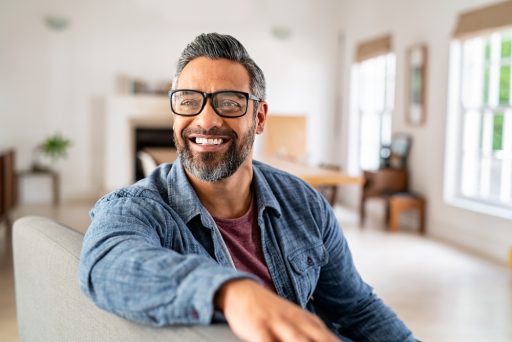 Smiling middle-aged man with glasses and a greying beard, sitting on a sofa at home