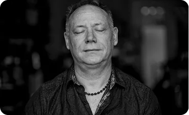 Victor Towle with eyes closed in a calm, meditative pose, wearing a beaded necklace and a dark patterned shirt, captured in black and white.