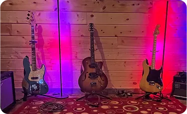 Three guitars resting on stands against a wooden wall, illuminated by colorful stage lighting in red, blue, and purple tones.