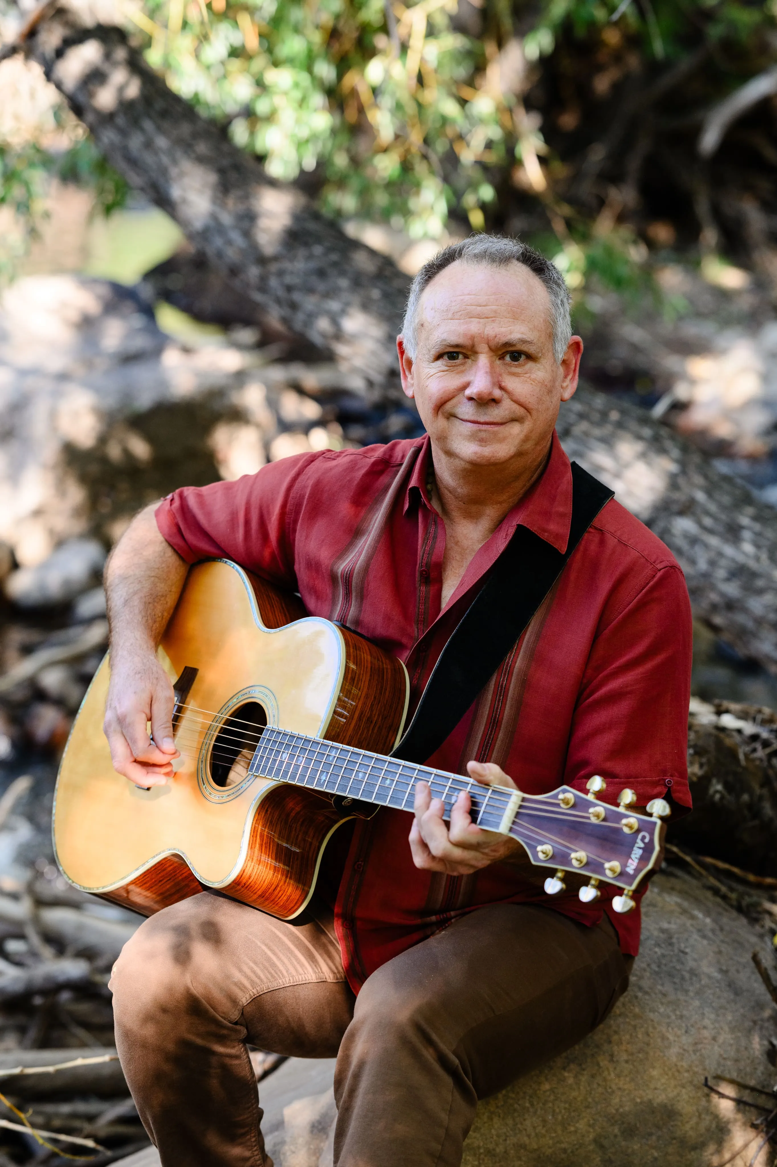 Victor Towle sitting outdoors on a rock, playing an acoustic guitar and looking at the camera, surrounded by trees and natural scenery.