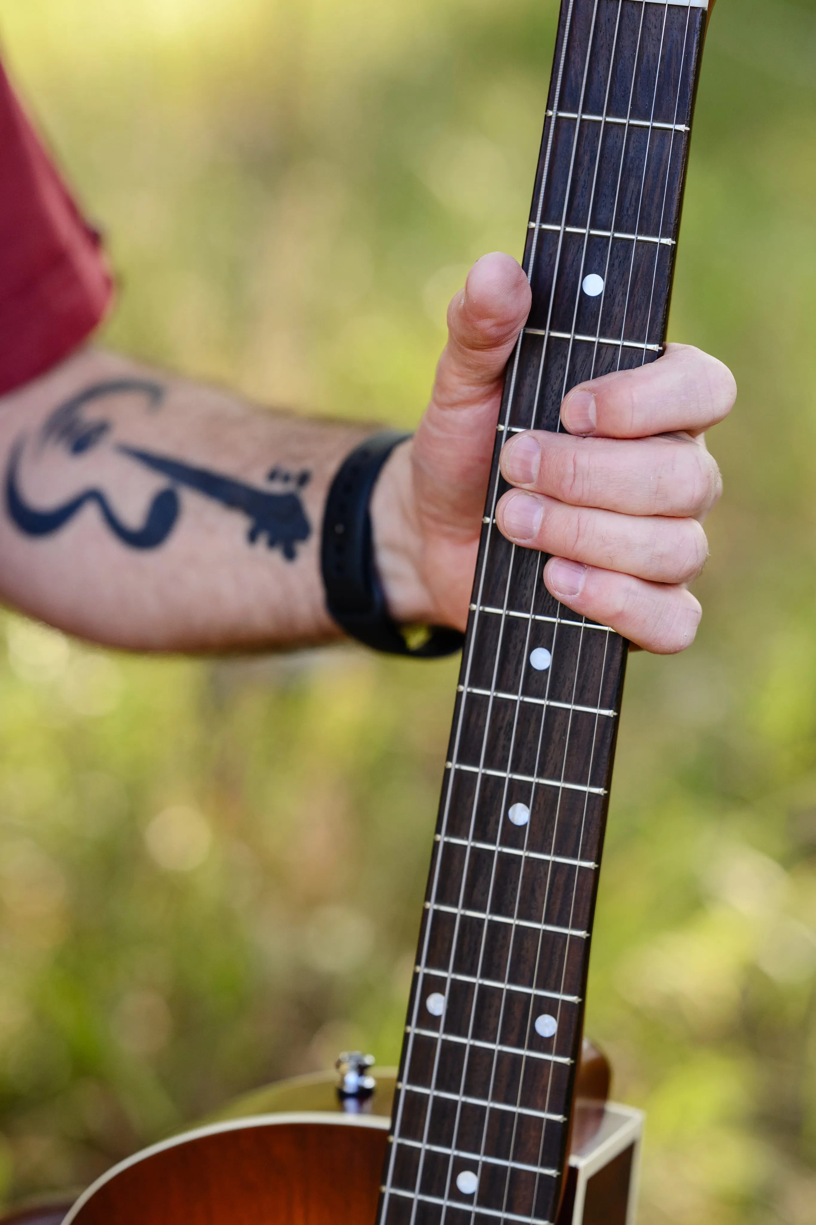 Close-up of a tattooed arm gripping the neck of an acoustic guitar, with a guitar silhouette tattoo visible and a blurred natural background.