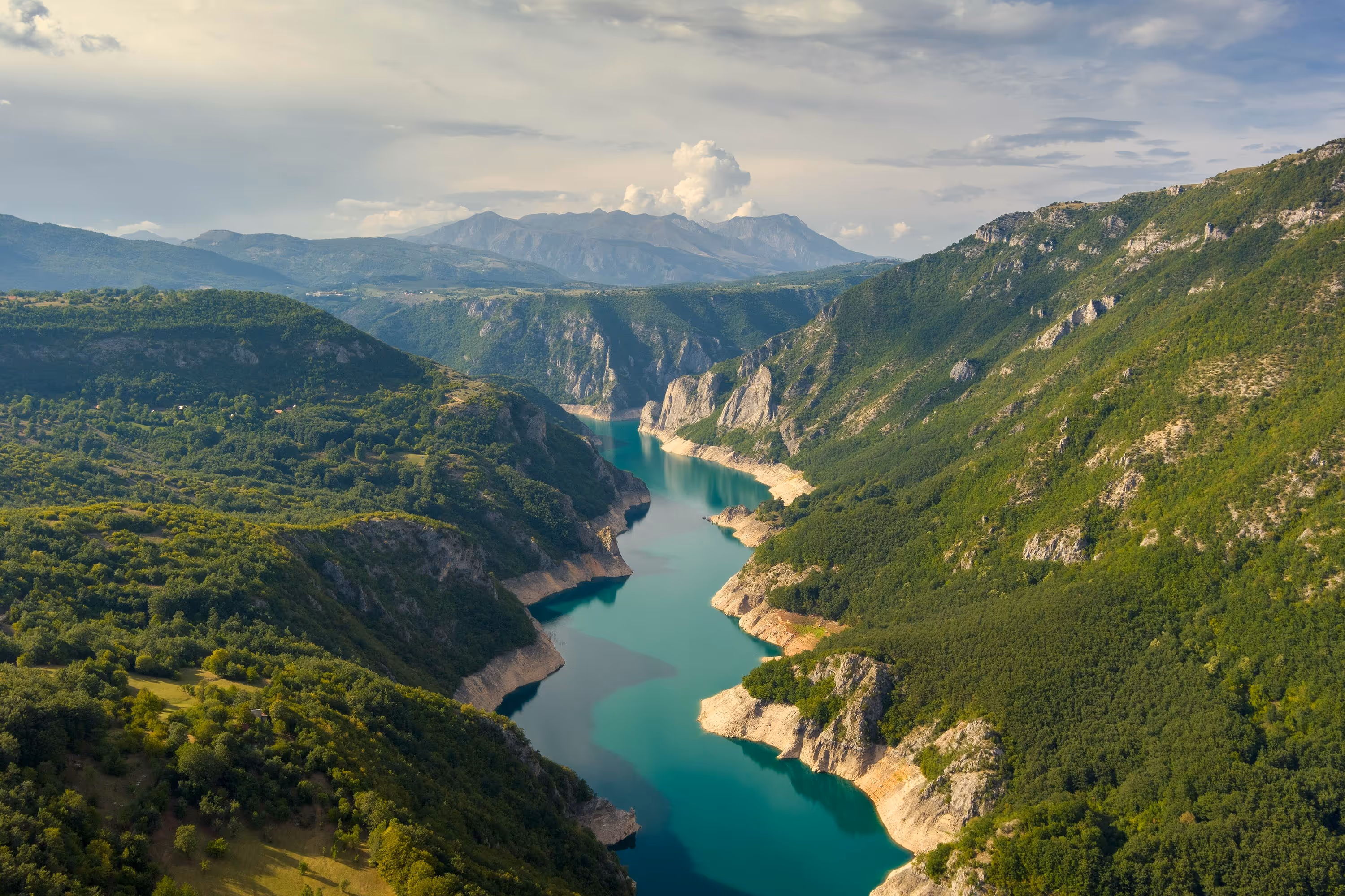 Aerial view of a turquoise river winding through a lush green mountain valley in Montenegro, with dramatic cliffs and distant peaks under a soft cloudy sky.