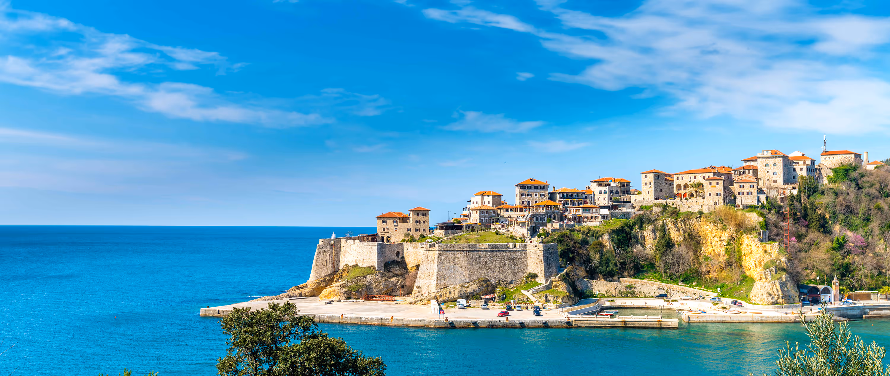 Scenic view of Ulcinj’s historic Old Town perched on a rocky cliff above the Adriatic Sea, with stone houses and orange rooftops under a bright blue sky.