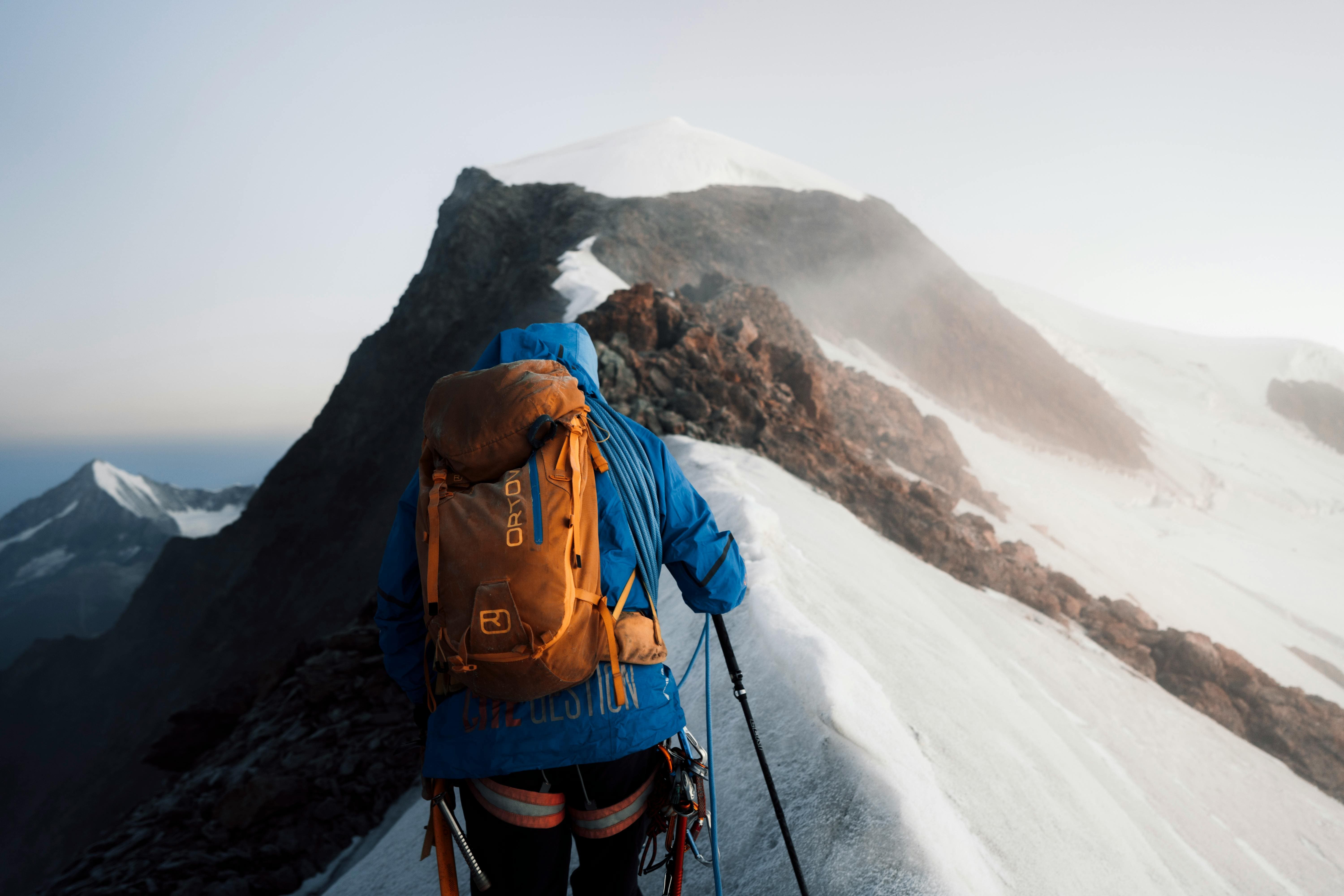 Trekkeur en veste bleue et sac à dos orange avançant sur une arête enneigée vers un sommet, au Népal.