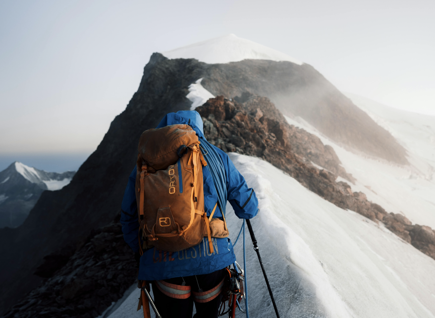Trekkeur en veste bleue et sac à dos orange avançant sur une arête enneigée vers un sommet, au Népal.