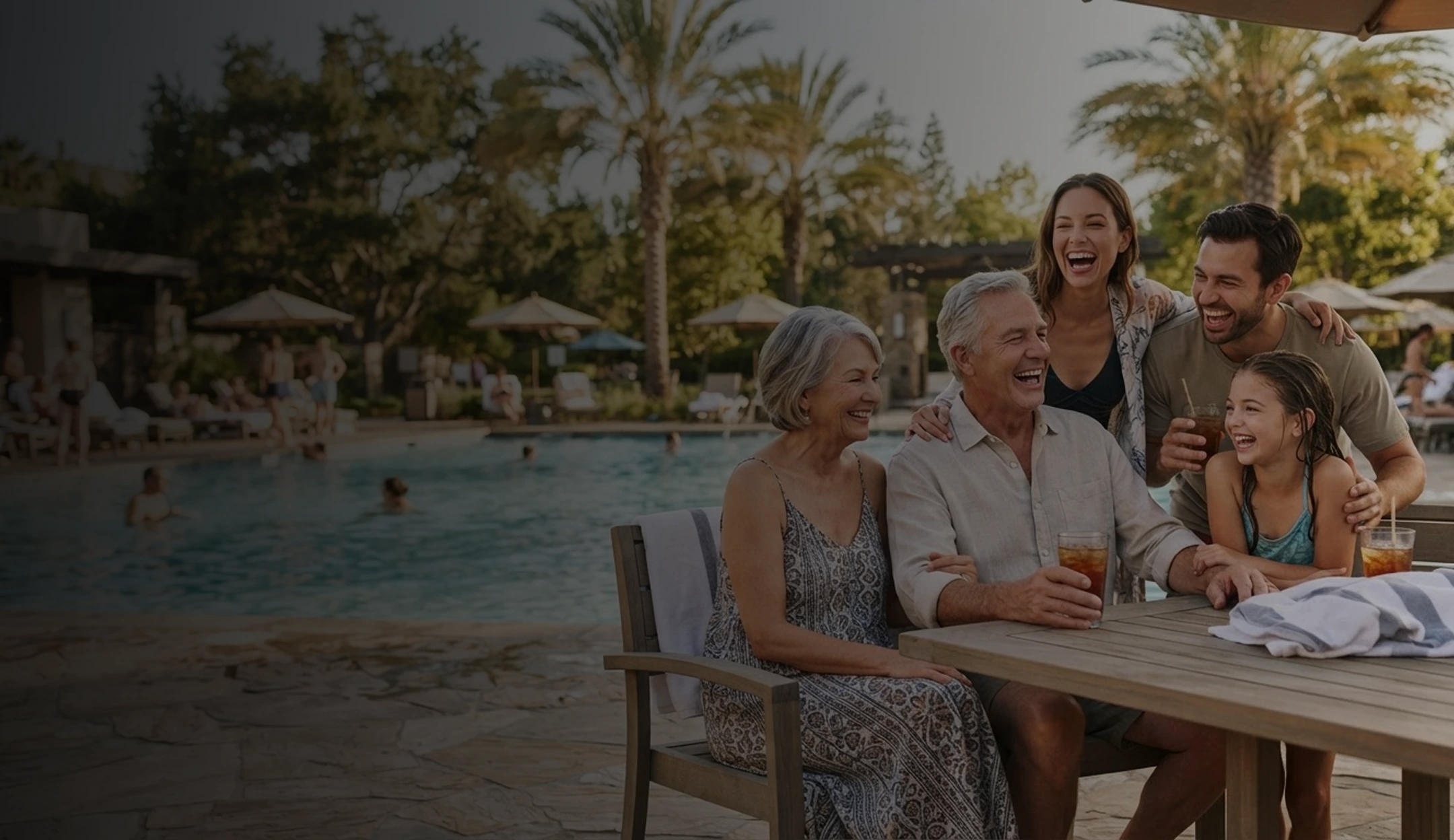Multigenerational family laughing and enjoying cold drinks while sitting at a wooden table by a swimming pool with palm trees and lounge chairs in the background.