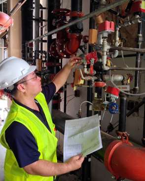 Worker wearing a white hard hat and neon yellow safety vest inspecting a sprinkler system valve while holding a checklist.