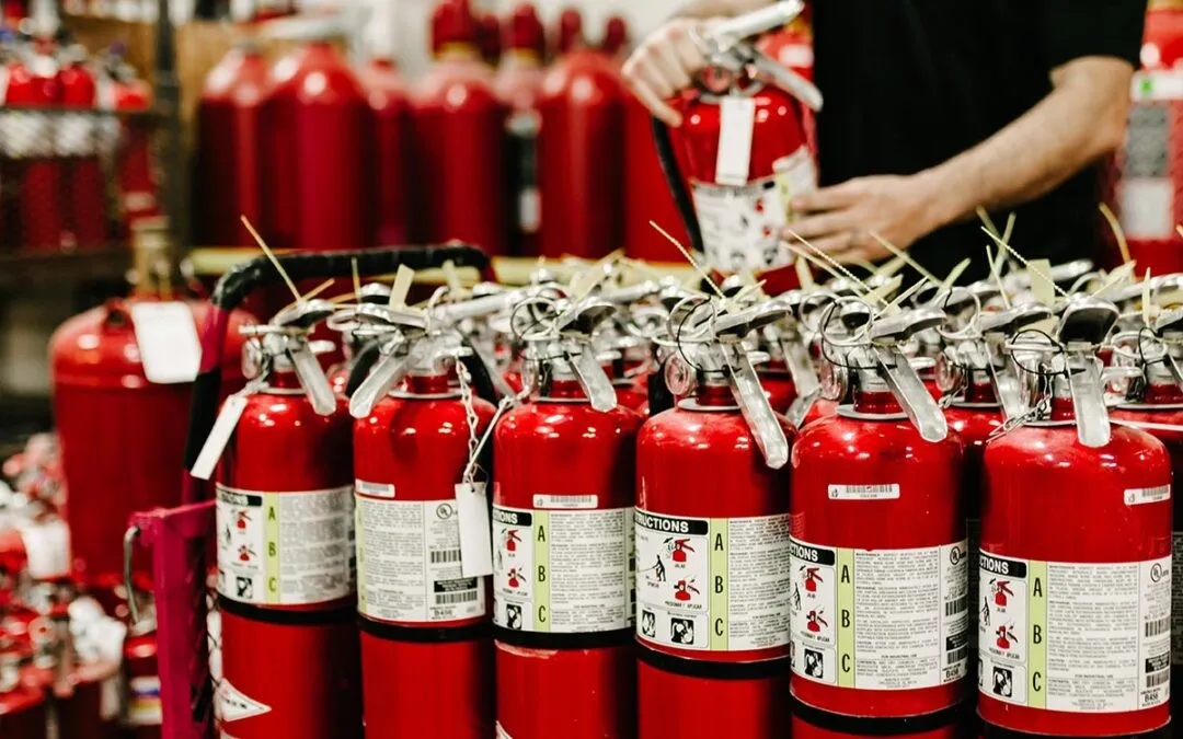 Rows of red fire extinguishers with labels in a storage area and a person handling one extinguisher in the background.