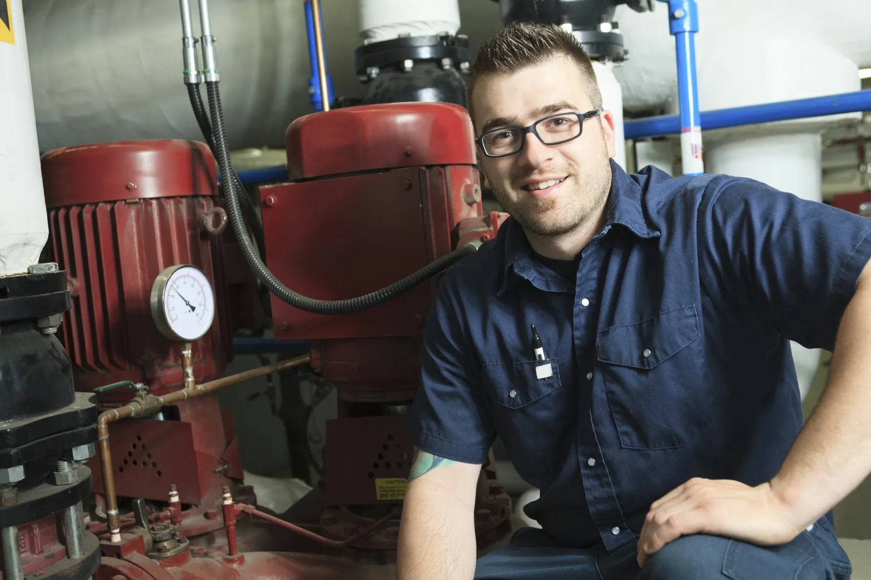 Smiling technician in blue work shirt with pen in pocket kneeling beside red industrial machinery with pressure gauge.