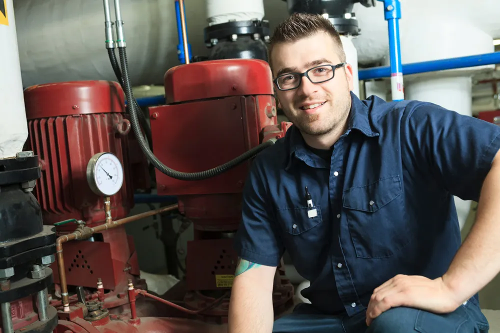 Smiling technician in glasses and navy blue work shirt leaning on machinery with pressure gauge in industrial setting.