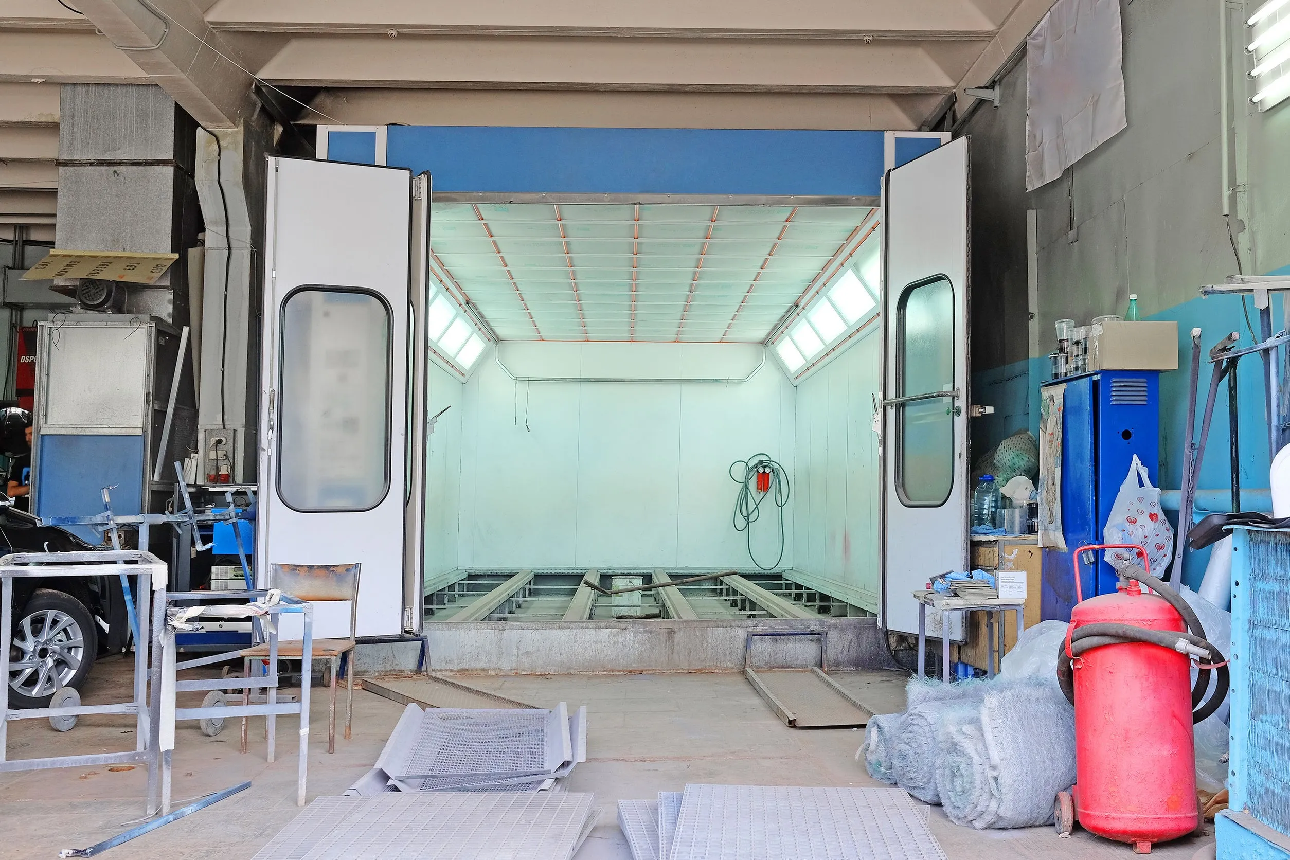 Empty industrial spray booth room with open double doors, surrounded by tools, chairs, and equipment.