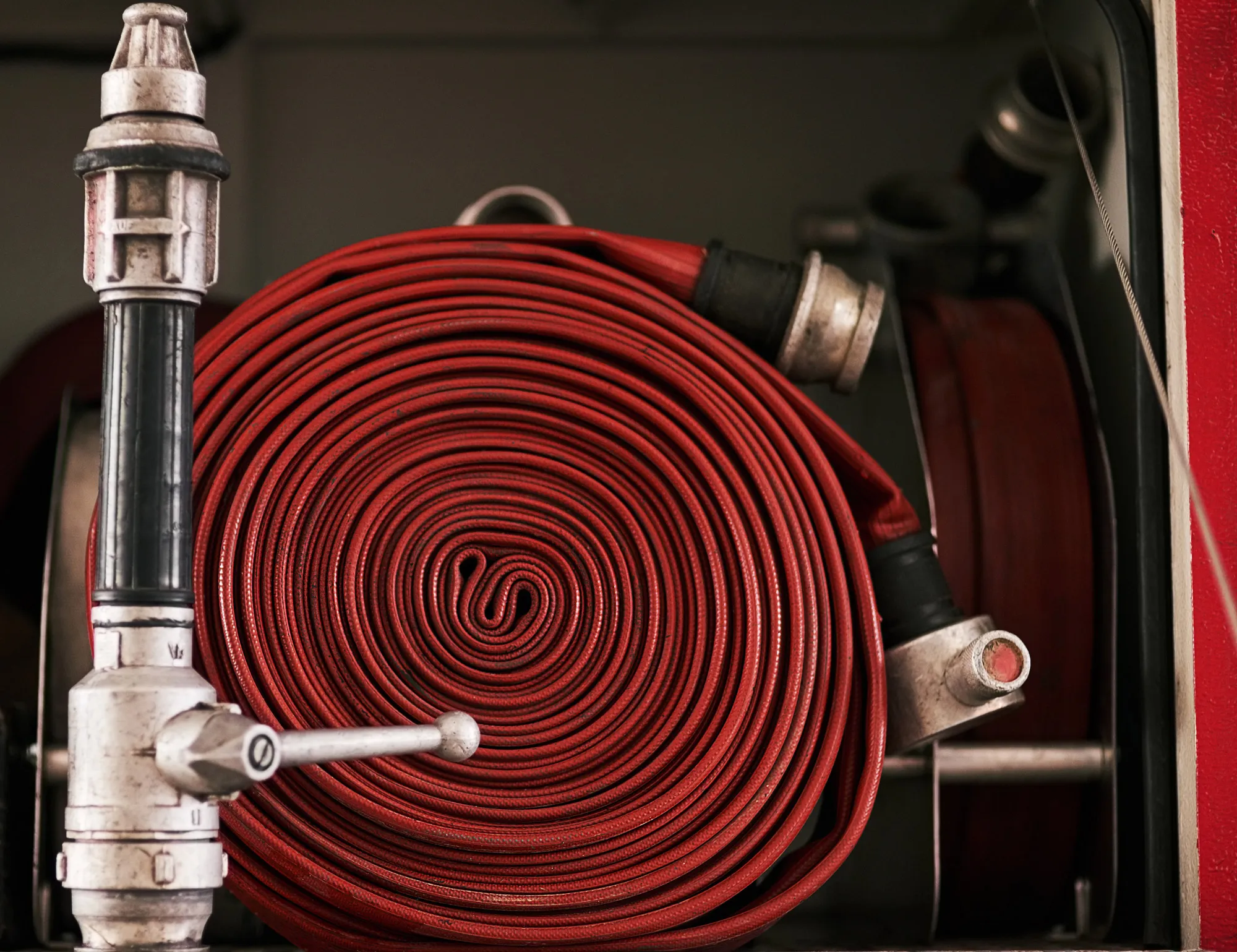 Coiled red fire hose with metal fittings mounted inside a fire truck compartment.