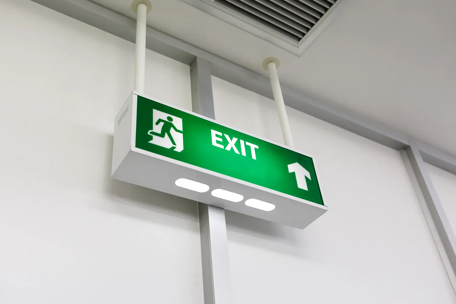 Green exit sign with a running figure and an arrow pointing up mounted on a white ceiling.