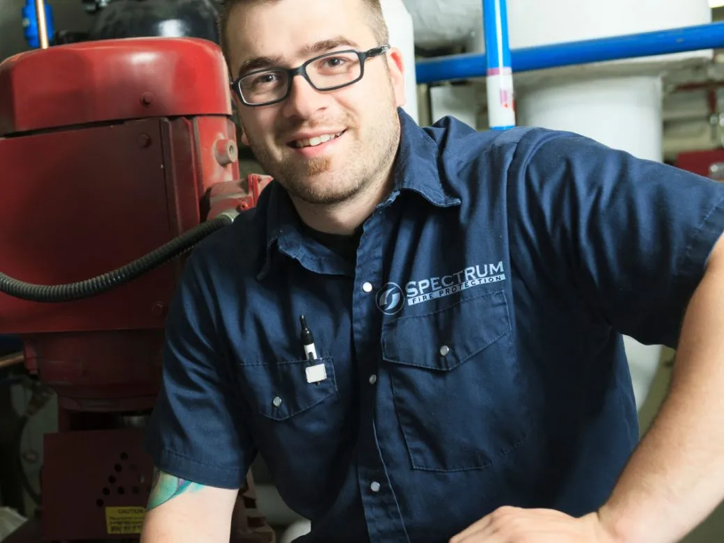 Man wearing glasses and a navy blue shirt with Spectrum Fire Protection logo smiling in an industrial setting.