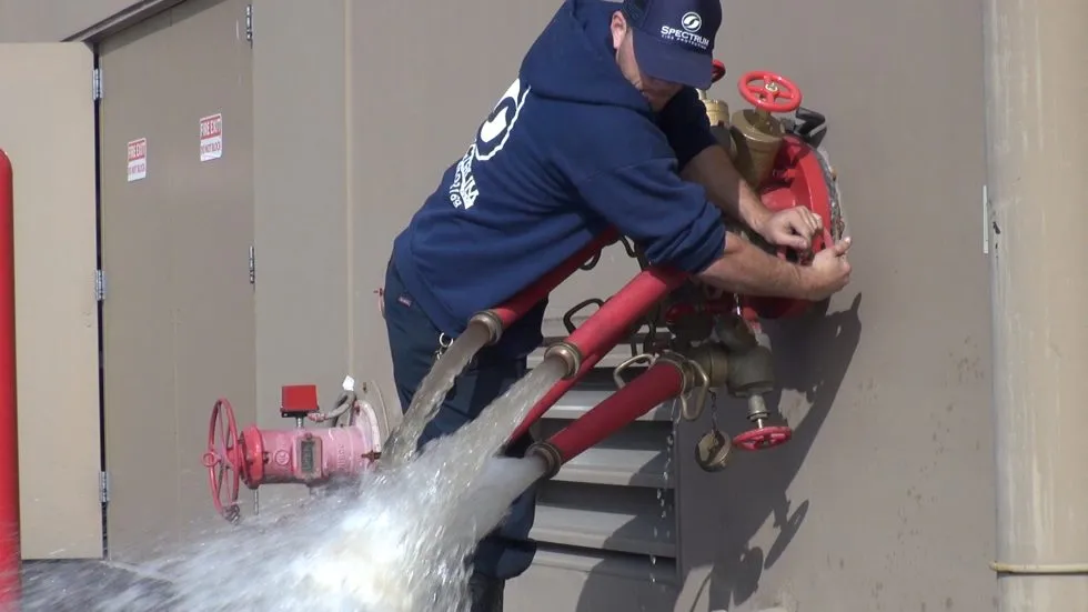 Man in a navy Blue Spectrum hoodie and cap operating a red fire hydrant valve with water forcefully flowing out through connected red pipes.