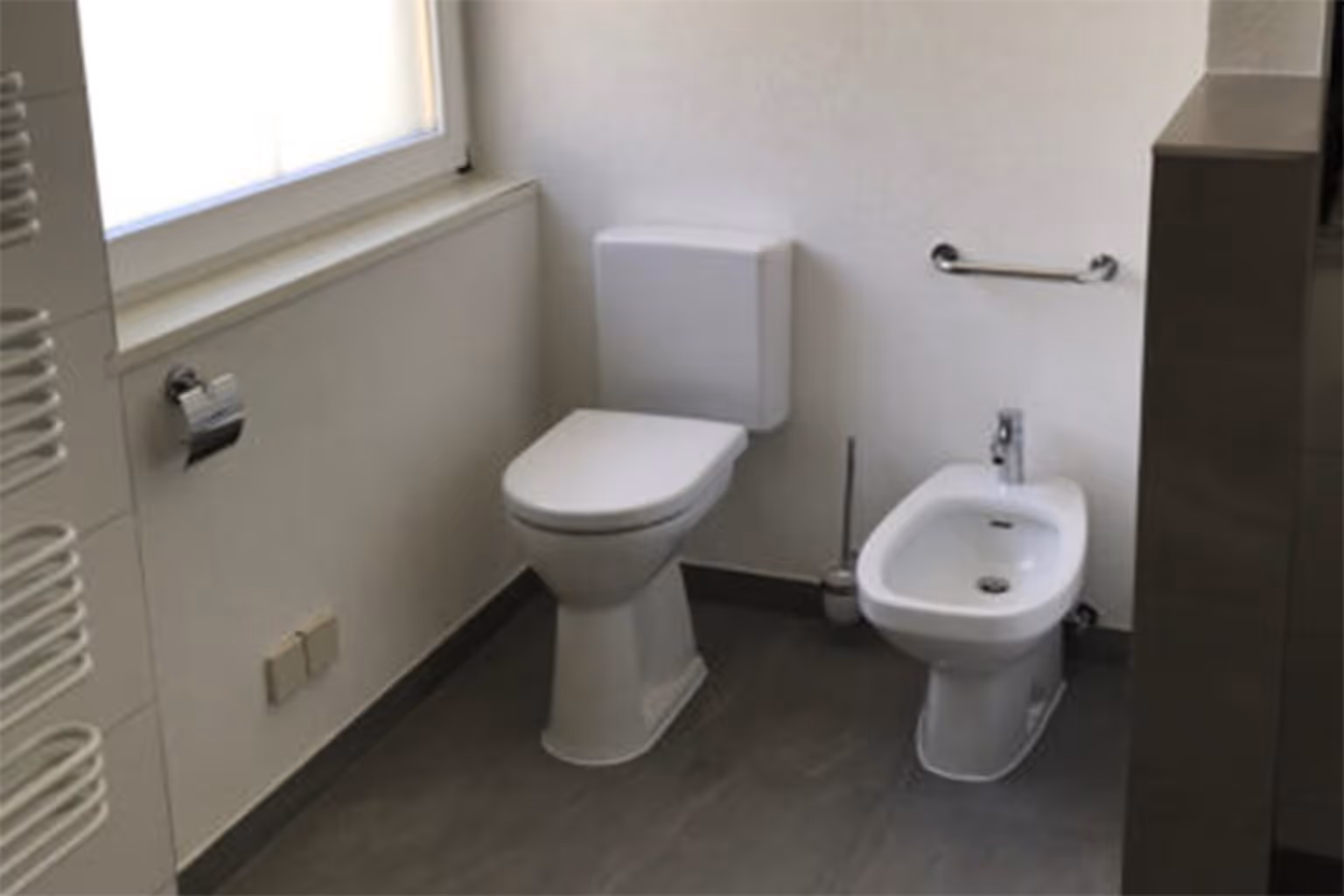 Unrenoviertes WC mit weißen Wänden und einfacher Keramikausstattung.