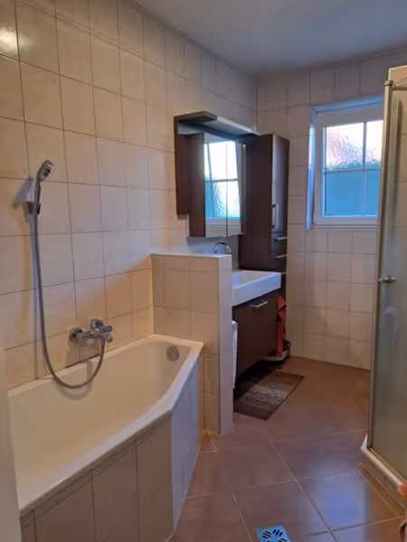 Altes Bad mit beigen Fliesen, Badewanne und kleinem Fenster vor der Sanierung.
