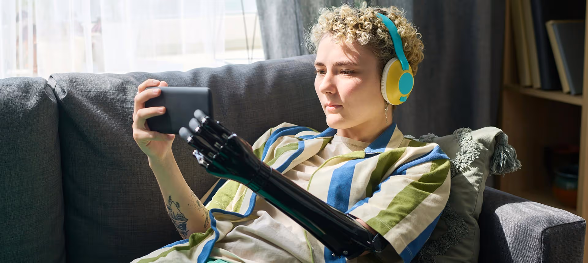 Eine Person mit Arm-Prothese entspannt mit bunten Kopfhörern und einem Tablet auf einem Sofa.