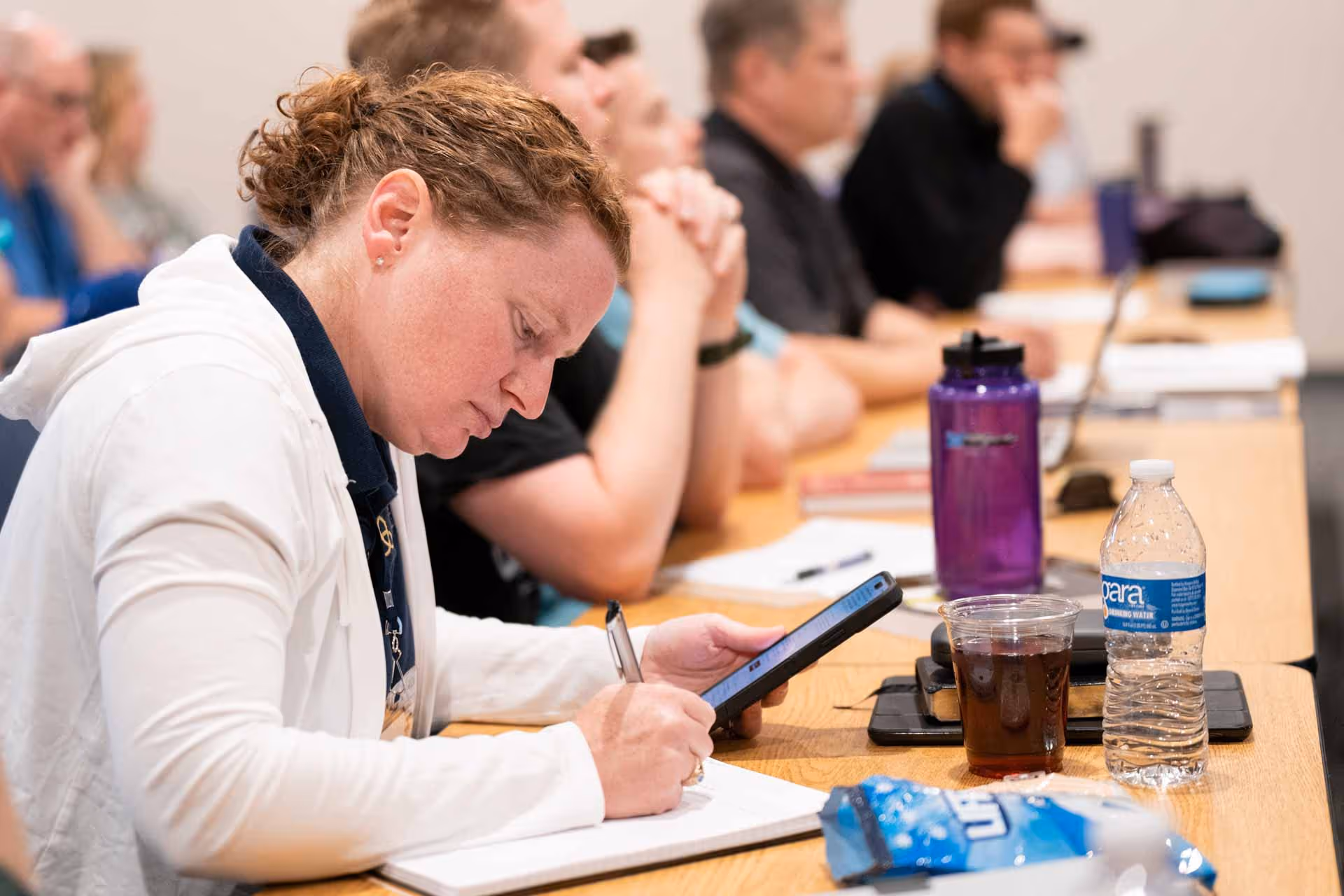 A woman with curly hair writes notes while looking at her phone in a classroom setting. Others around her listen attentively, conveying focus.