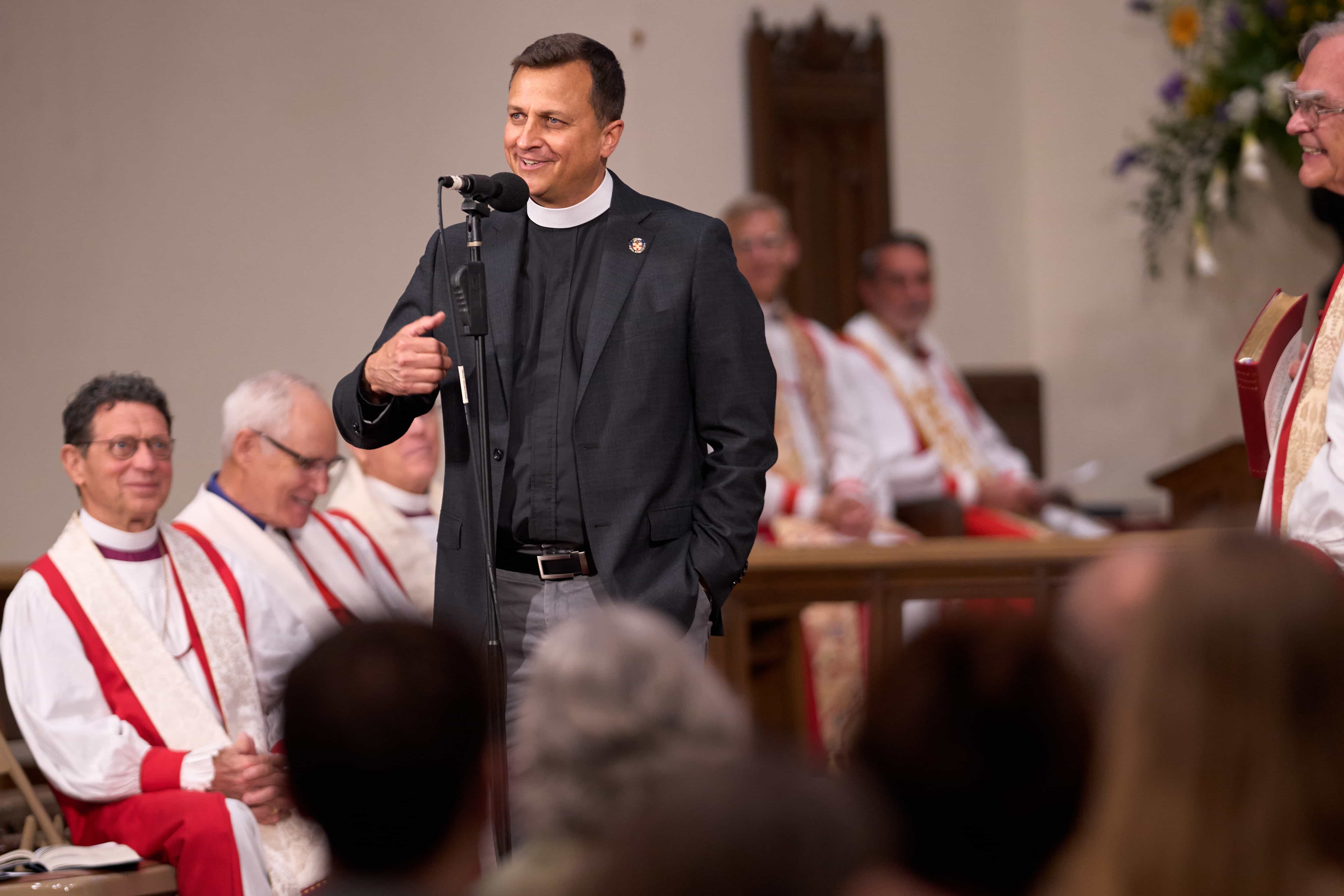 A priest smiles while speaking at a microphone in a church. Seated bishops in white and red vestments listen, and attendees are visible in the foreground.