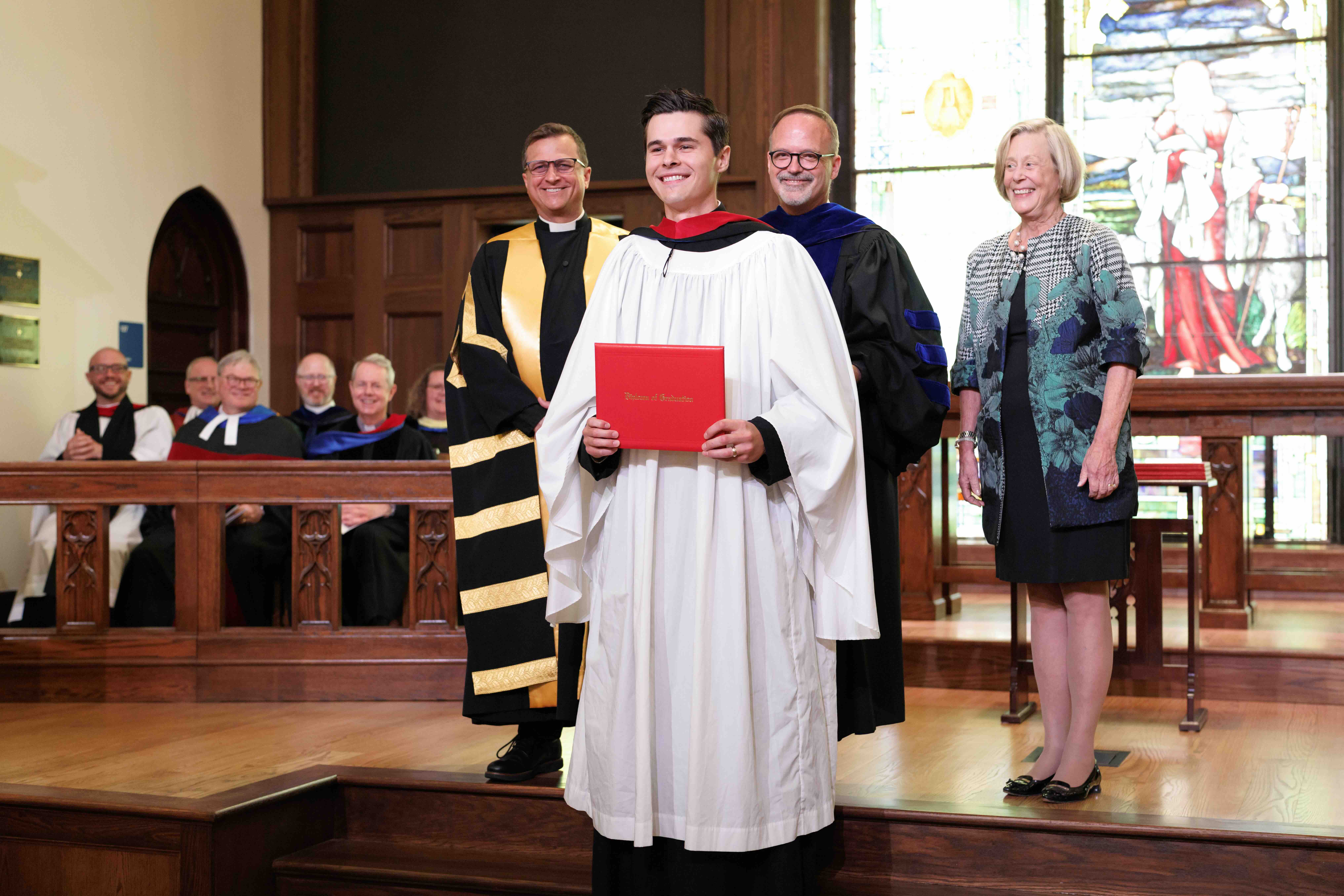 Smiling graduate in white robe holding a red diploma cover stands with three smiling faculty members in academic gowns inside a chapel with stained glass windows.