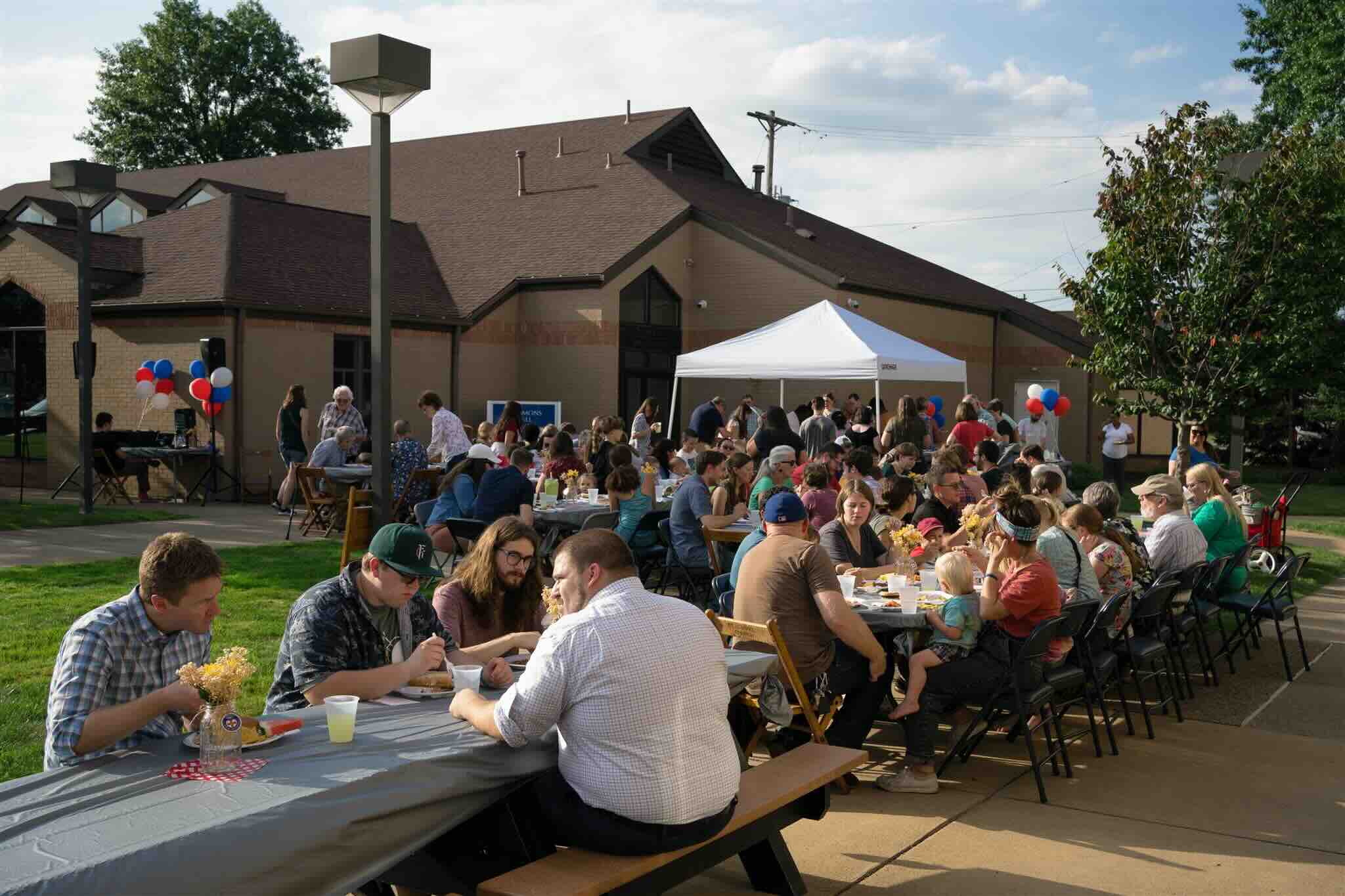 Outdoor community gathering with people seated at long tables, enjoying food beside a building. Decorations include red, white, and blue balloons.