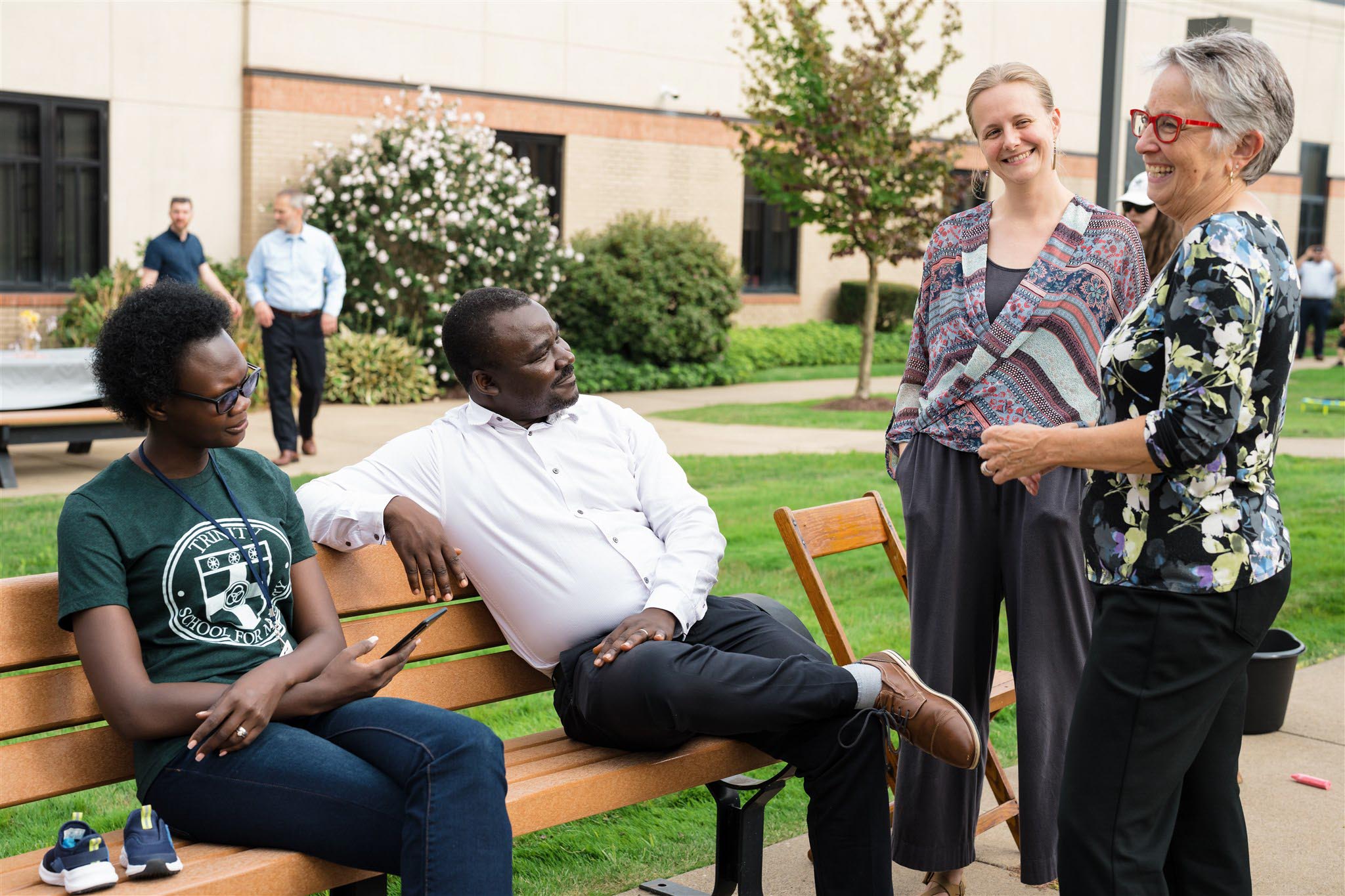 A group of four people enjoys a friendly conversation outdoors. Two sit on a bench while two stand nearby, all smiling. The background shows a garden and building.
