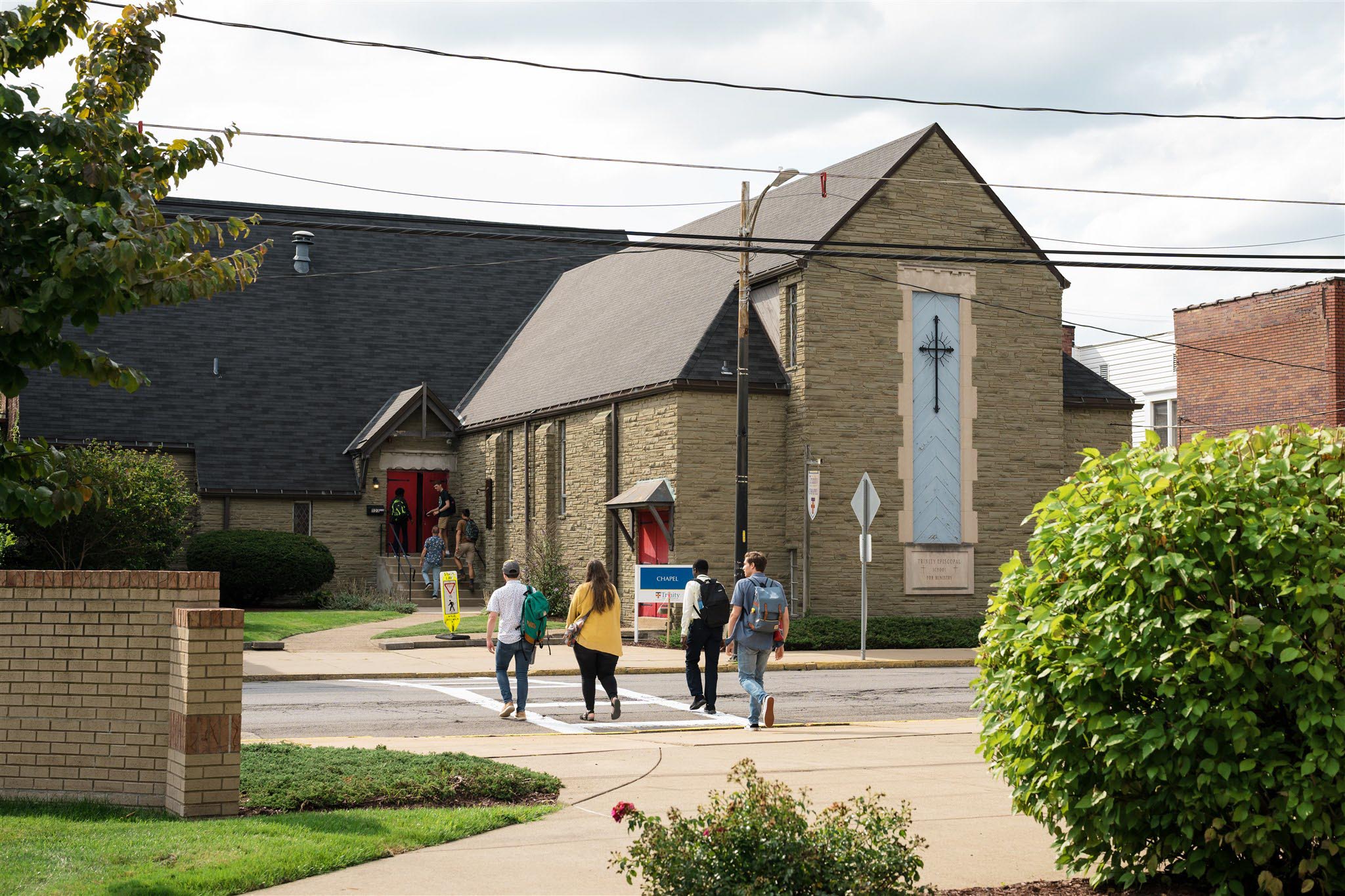 Group of people walking towards a stone church with a large cross on the facade. The atmosphere is sunny and relaxed, with greenery and clear skies.