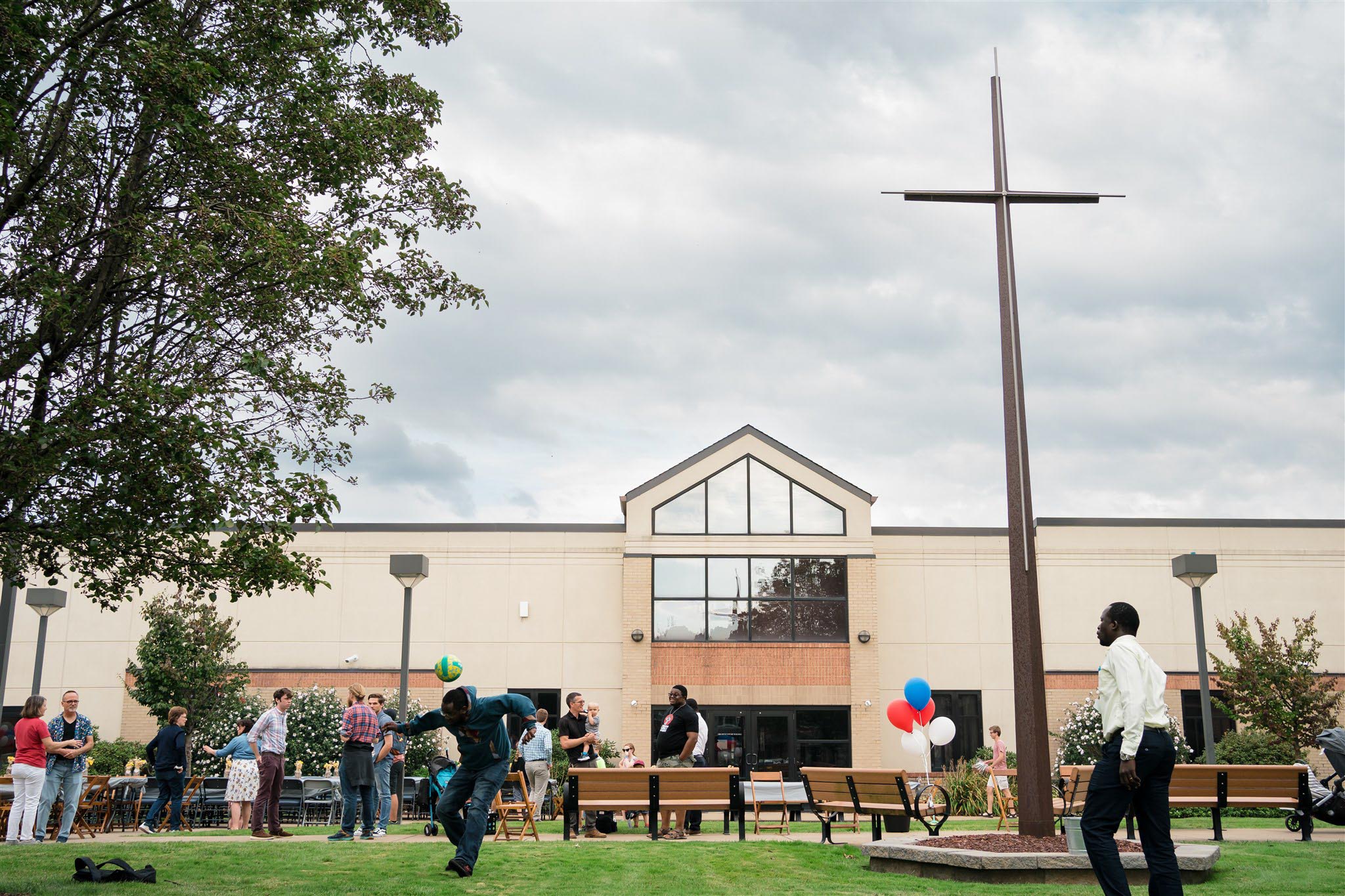 A group of people gathers outdoors near a large seminary building with a prominent cross. Balloons and lively interactions suggest a joyful event.