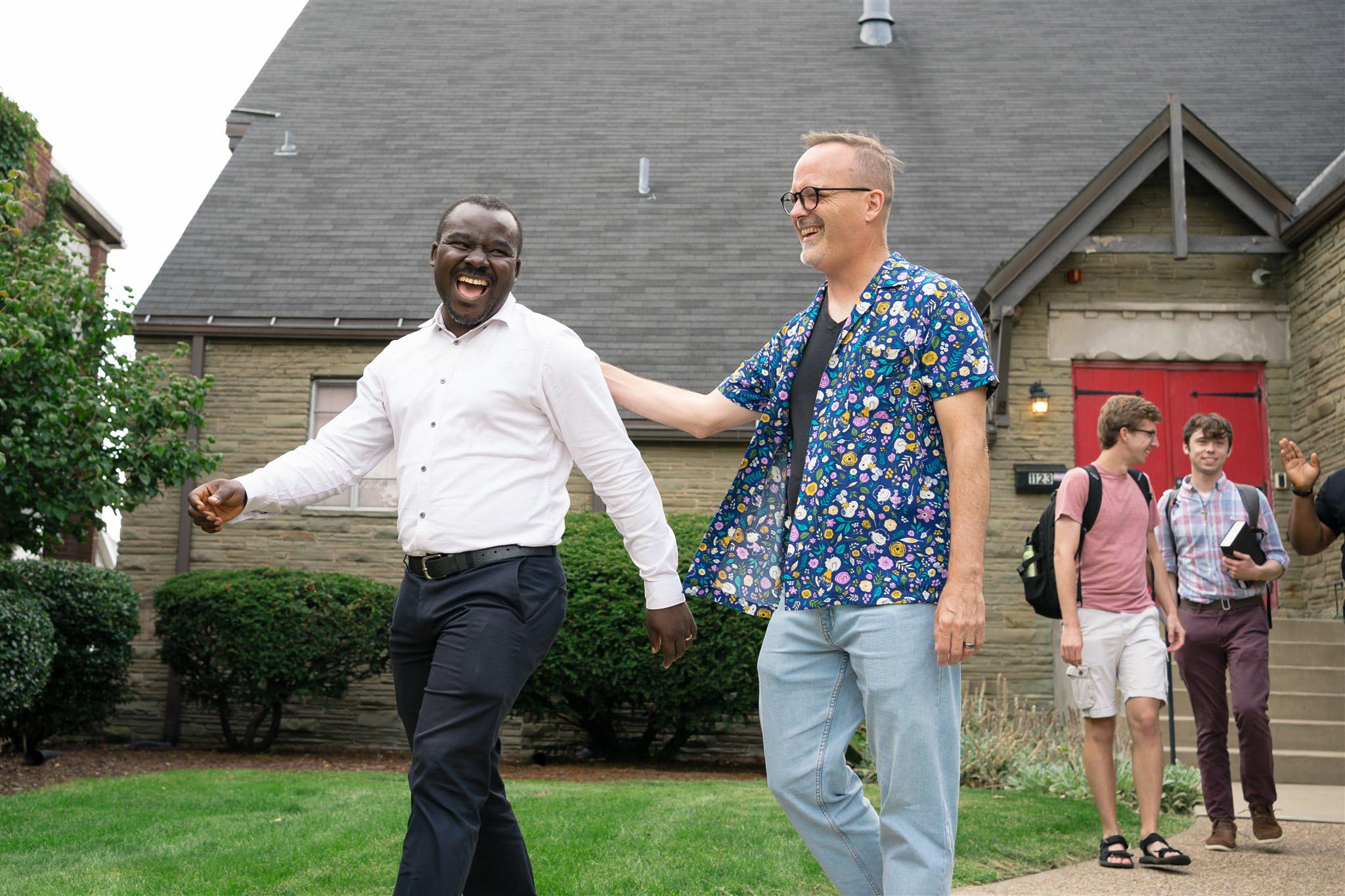 Two men share a joyful moment outside, one in a white shirt and the other in a floral shirt. Three young men converse in the background. Festive and lively atmosphere.