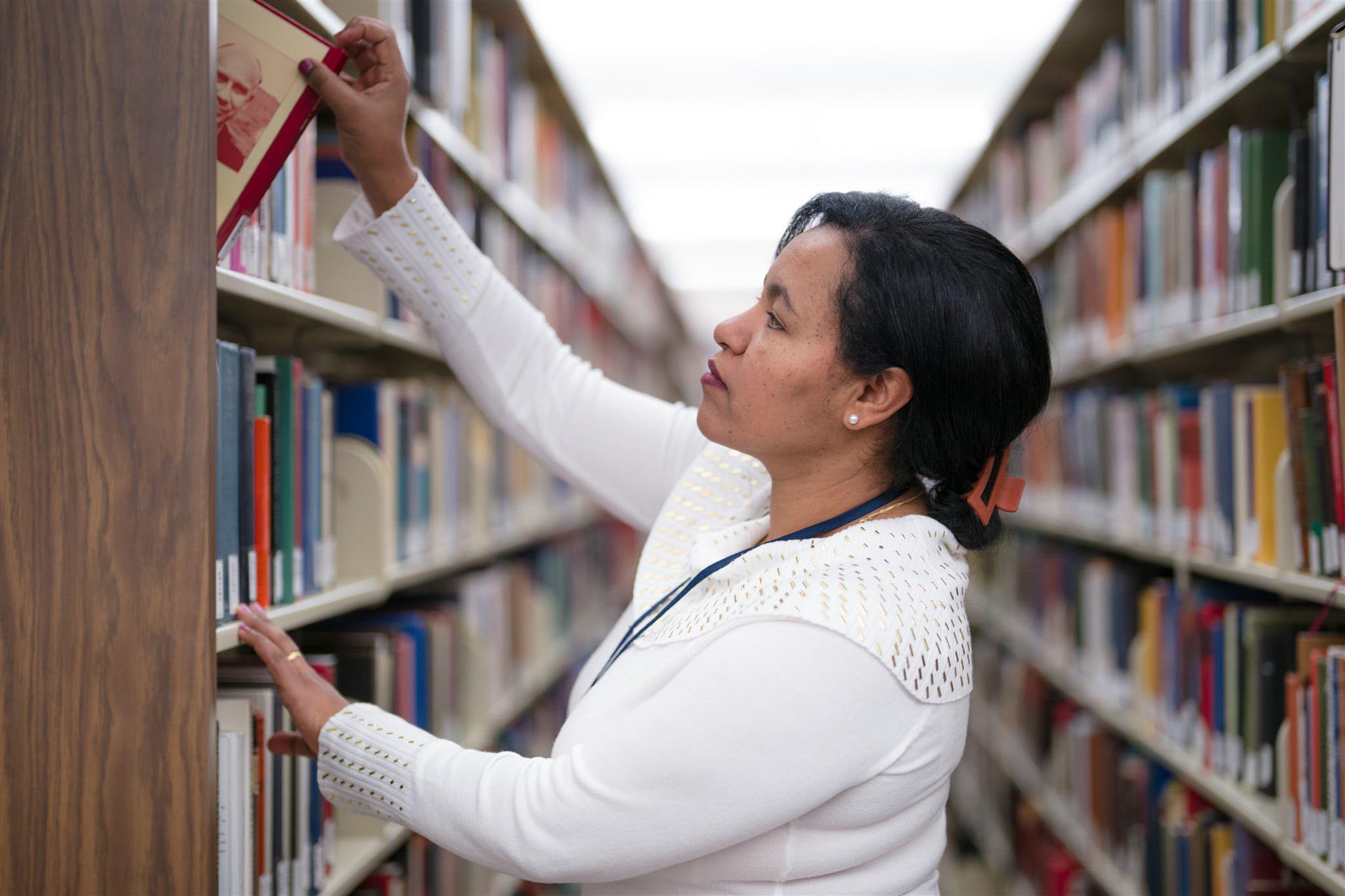 A woman in a white sweater reaches for a book on a tall library shelf. The aisle is lined with colorful books, creating a focused, studious atmosphere.