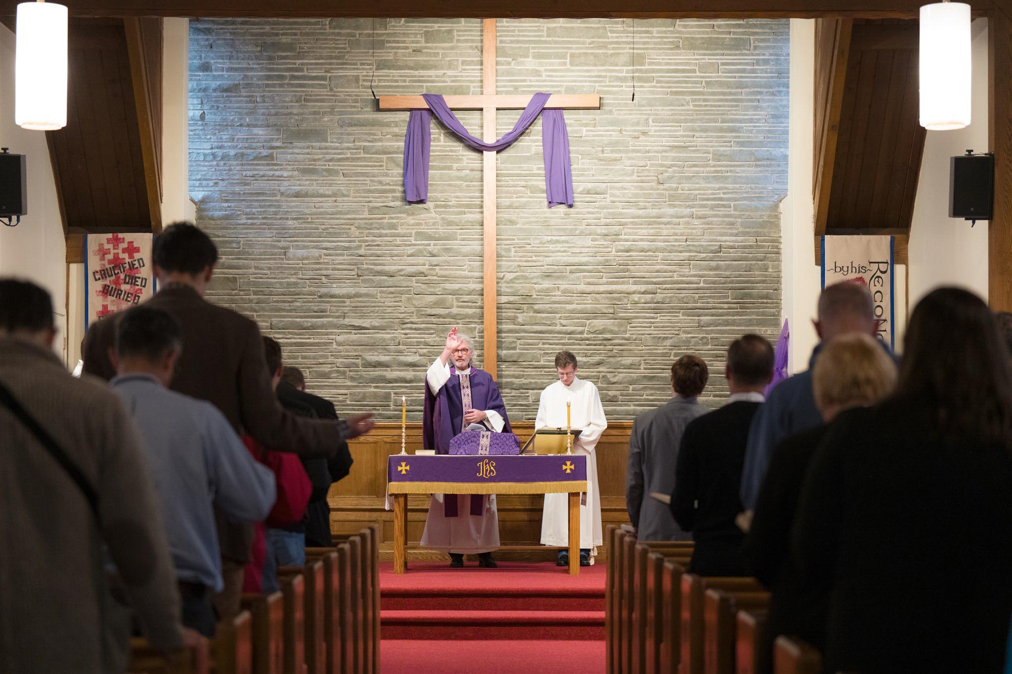 A priest and altar server conduct a service in a church, standing before a large cross draped in purple. Congregants are seated, facing them.