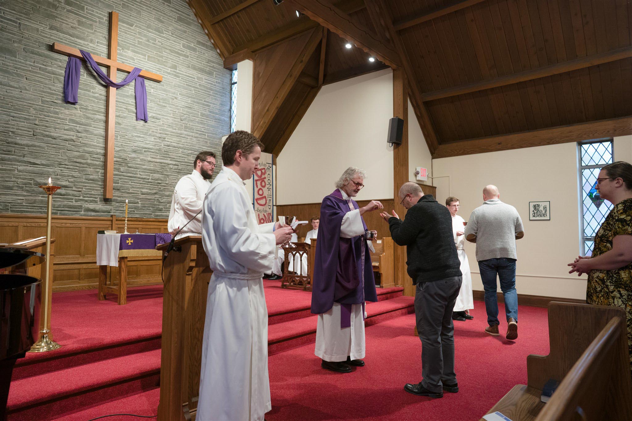 A priest in a purple robe conducts communion in a church with a wooden cross and red carpet. Congregants and assistants participate reverently.