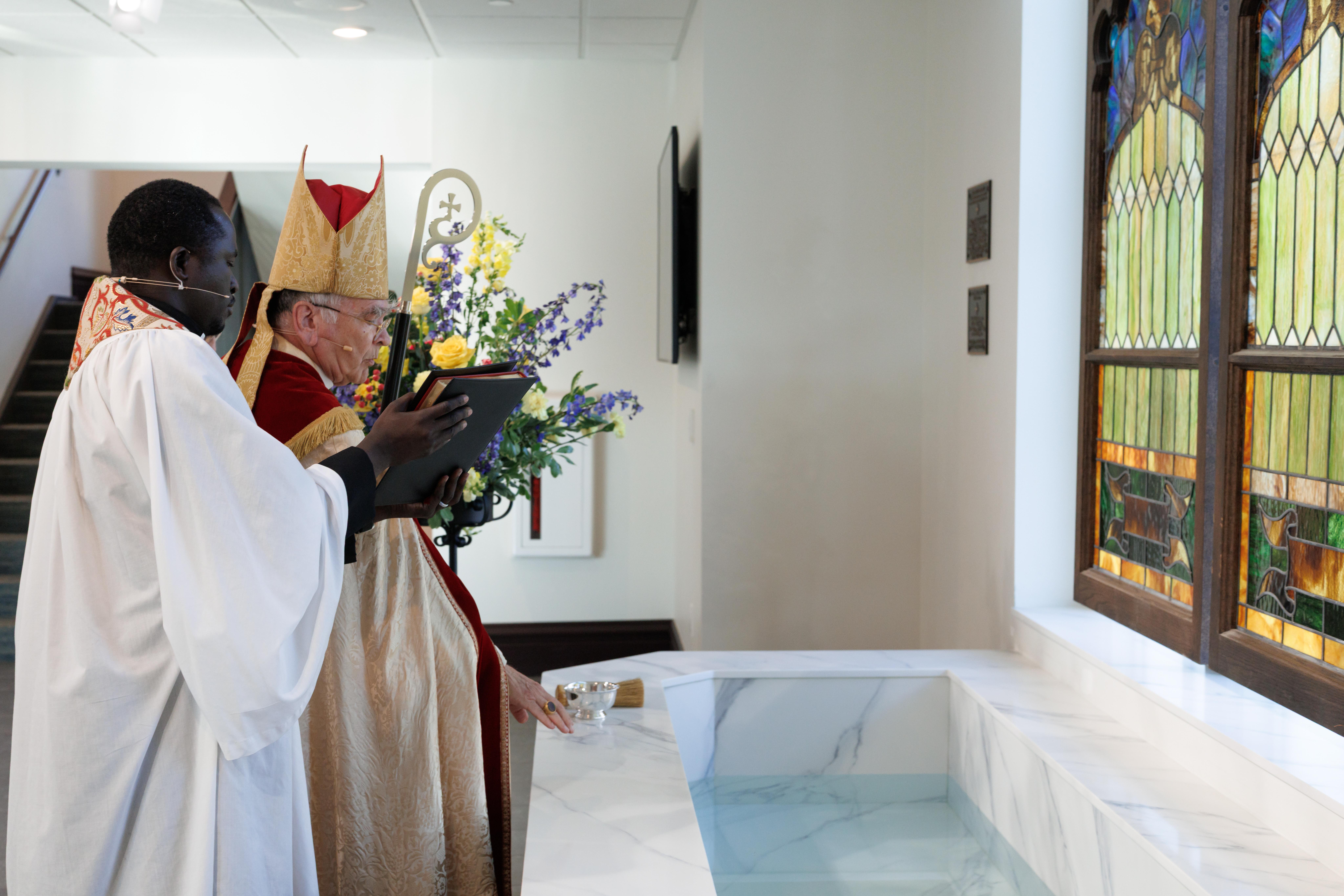 Two clergy members in ceremonial attire stand by a marble baptismal font. One holds a tablet, while colorful stained glass windows and flowers adorn the background, creating a solemn and reverent atmosphere.