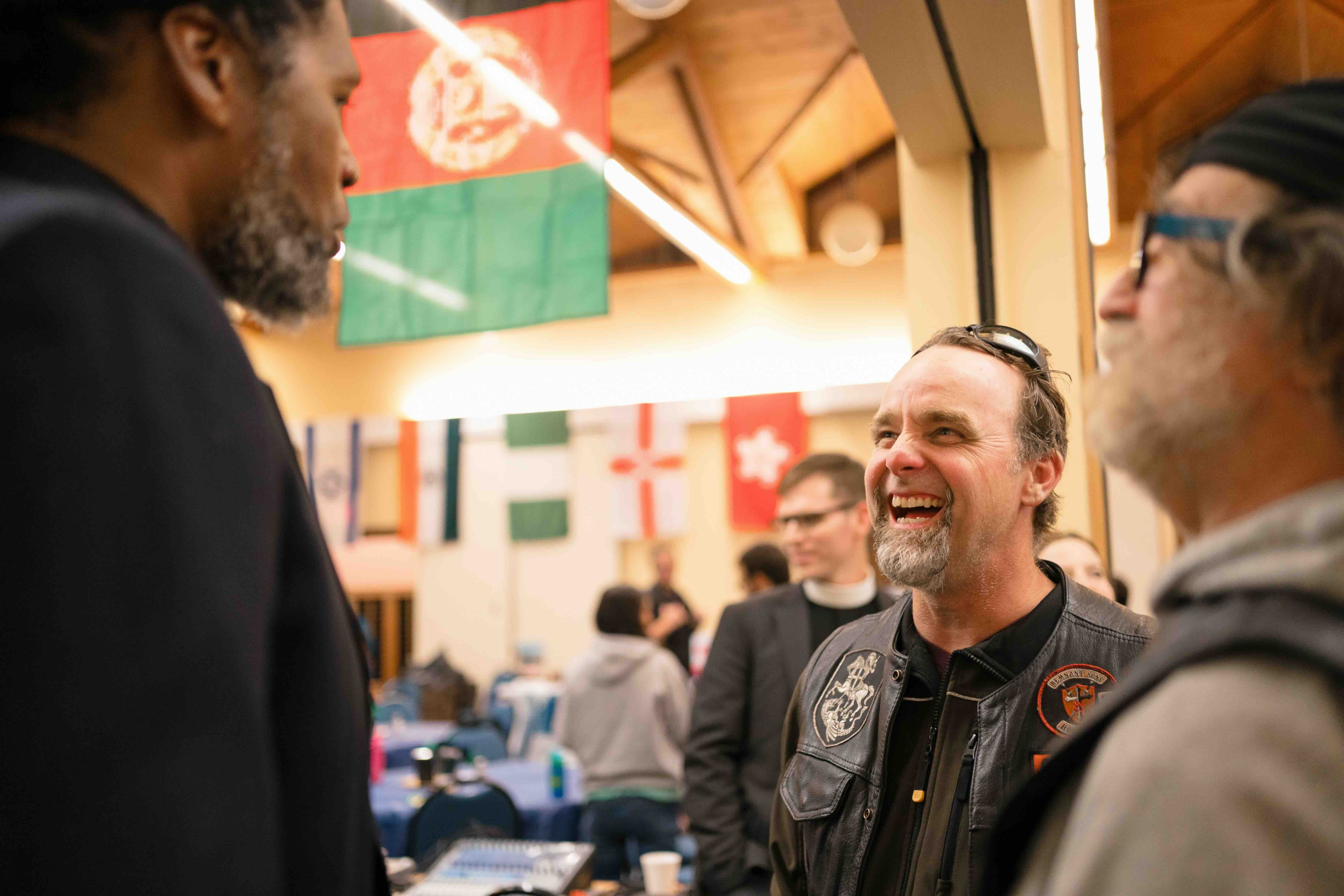 Three people in a lively conversation at an indoor event, surrounded by international flags. One person laughs heartily, creating a warm atmosphere.