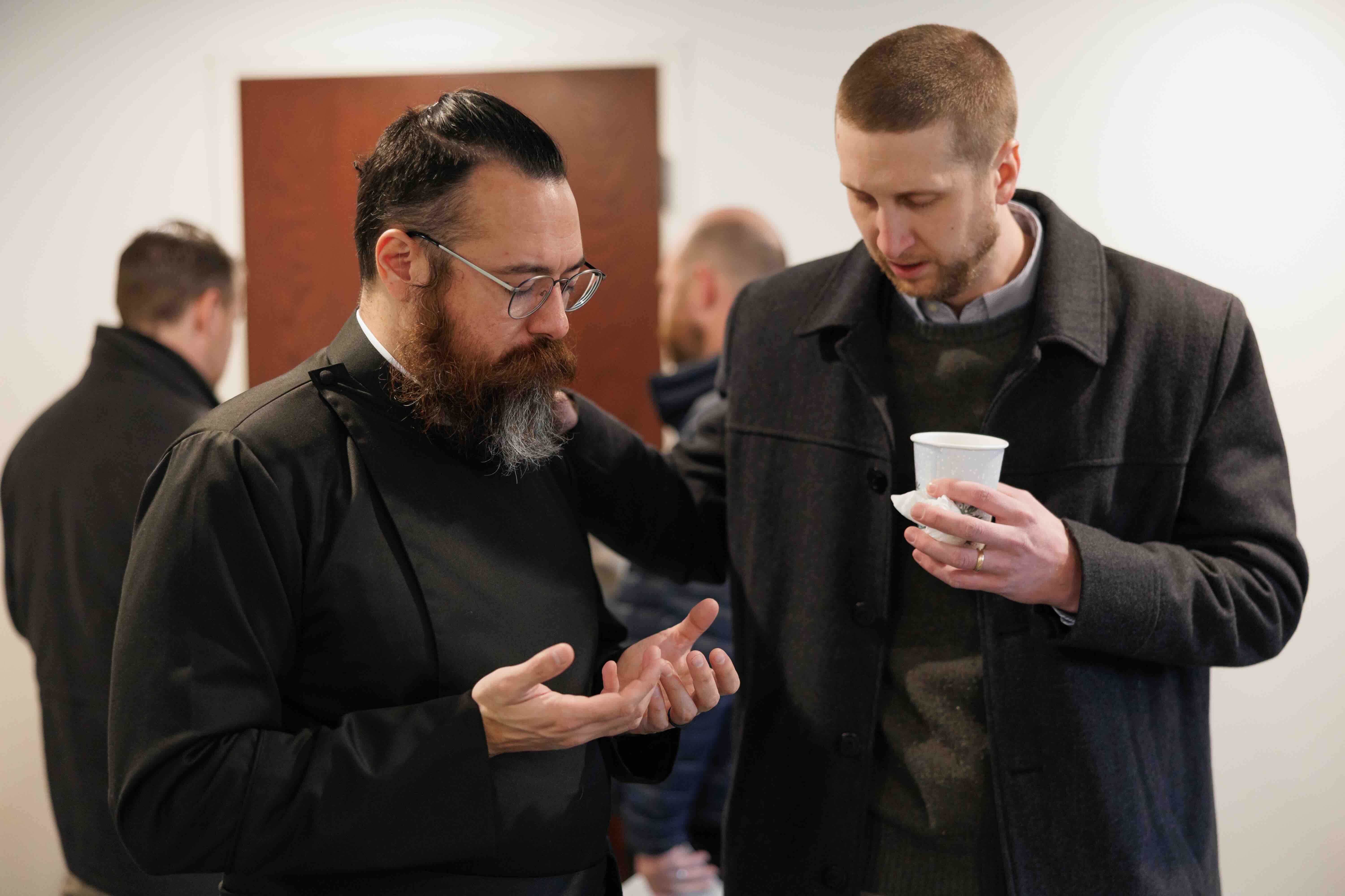 Two men engaged in a serious conversation, one wearing religious attire with glasses and a beard, the other holding a paper cup.