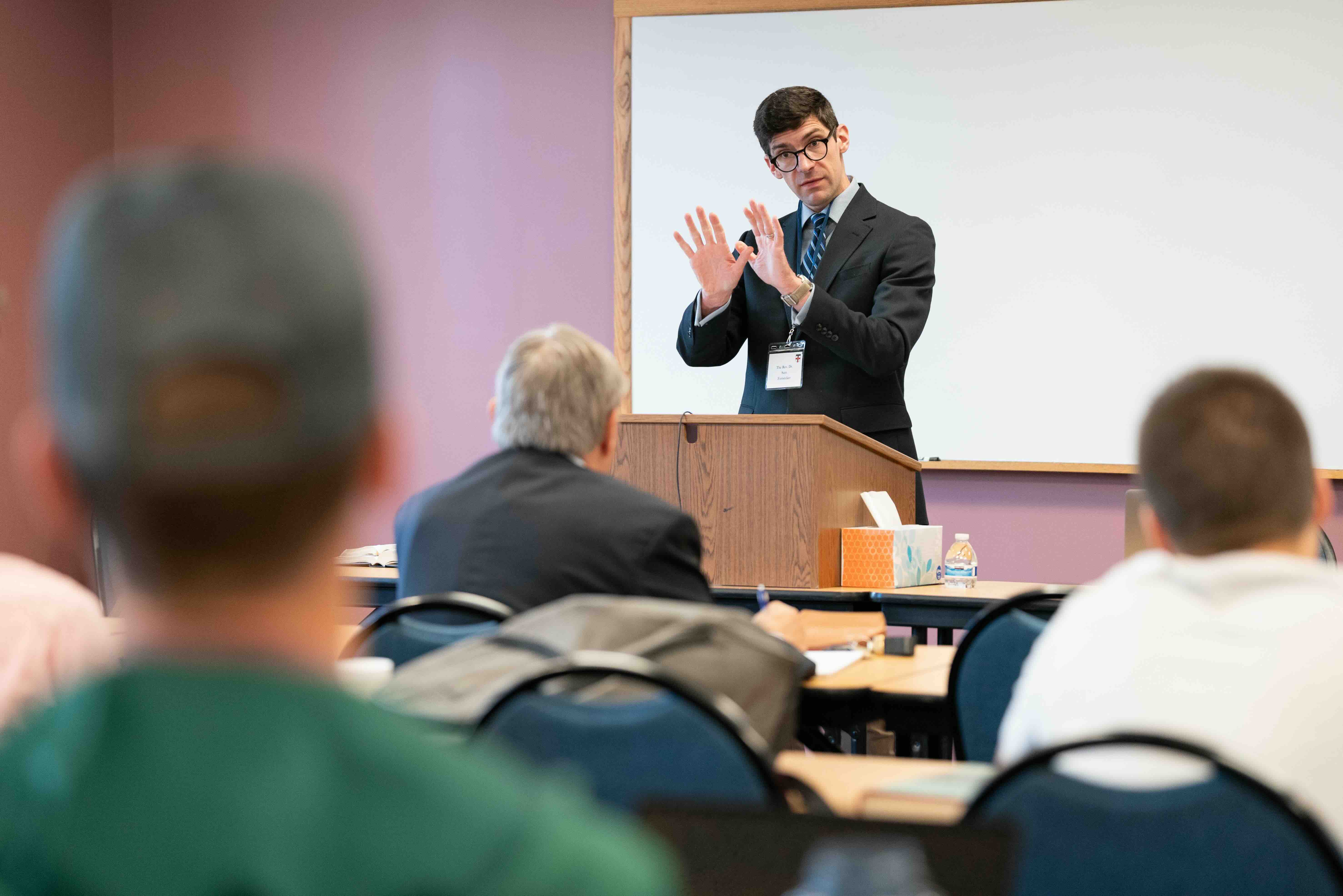 A man in a suit stands at a lectern, gesturing with his hands during a presentation. Several attendees face him, conveying an academic setting.