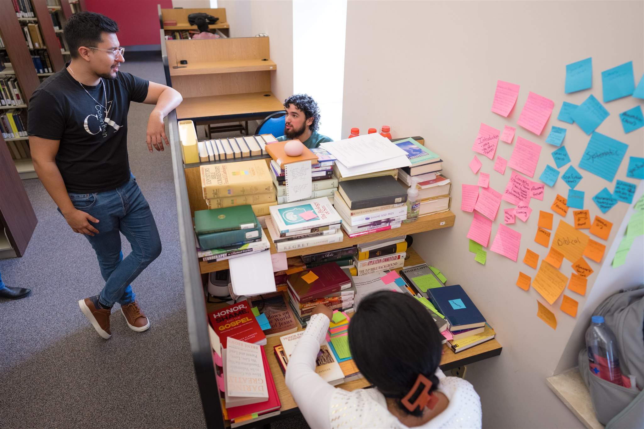 Three people are in a library study nook. One stands chatting, another sits behind a book-filled desk, and a third is writing. Walls are covered in colorful sticky notes.