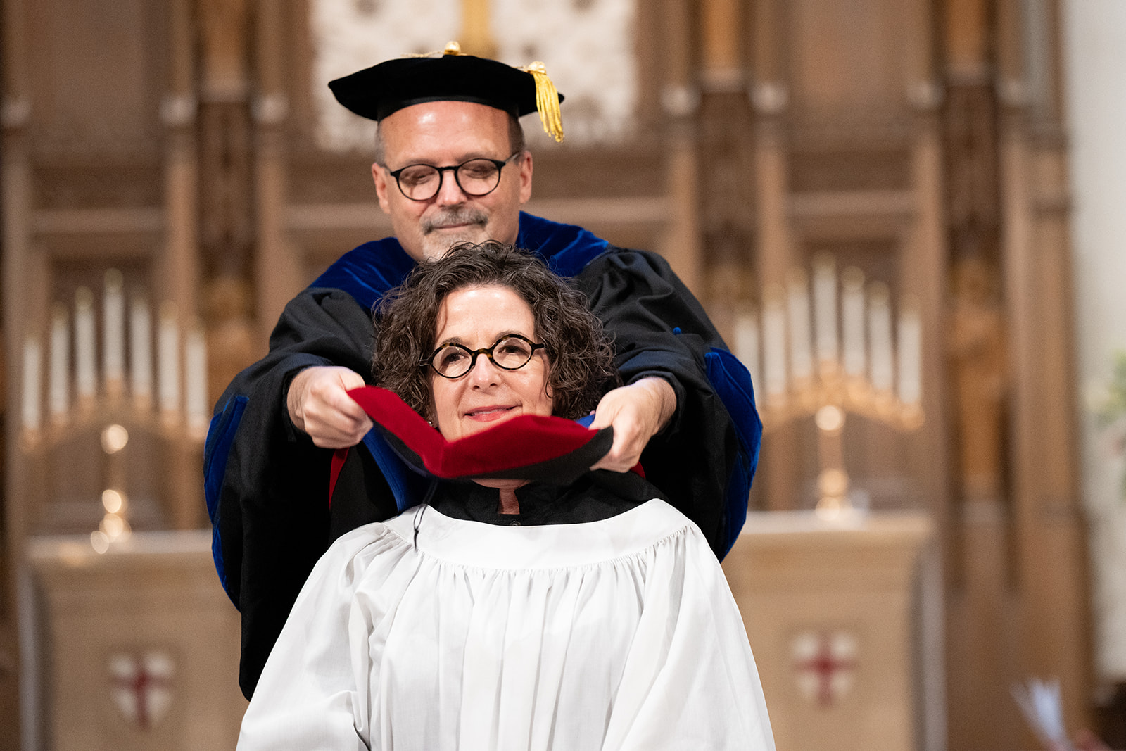Man in academic regalia placing a red hood on a woman in white graduation gown during a commencement ceremony.