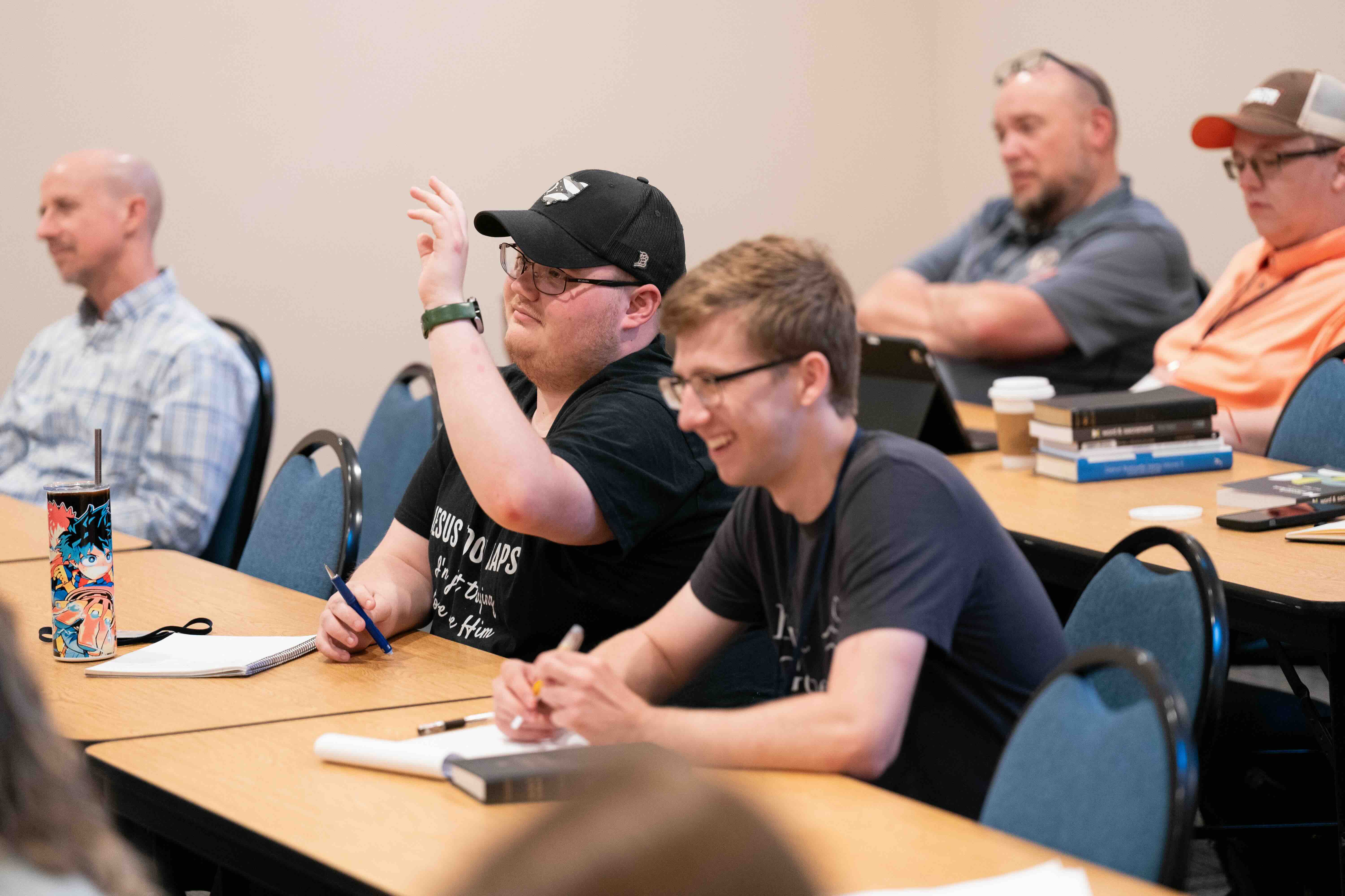 Students sitting in a classroom, one raising his hand while others listen and take notes.