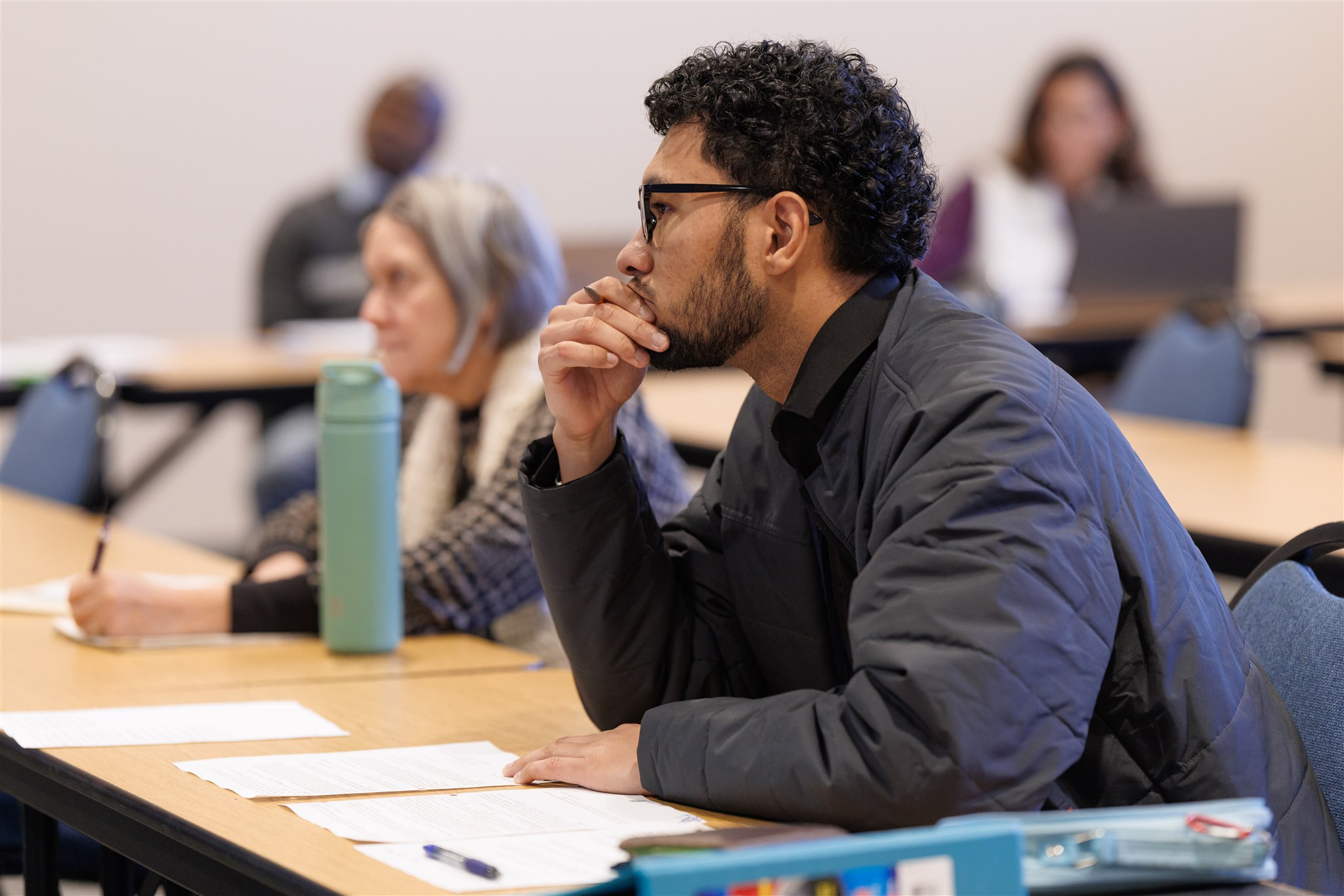 A man in a classroom setting attentively listens, resting his chin on his hand. Papers and a water bottle are on the table; others write notes.