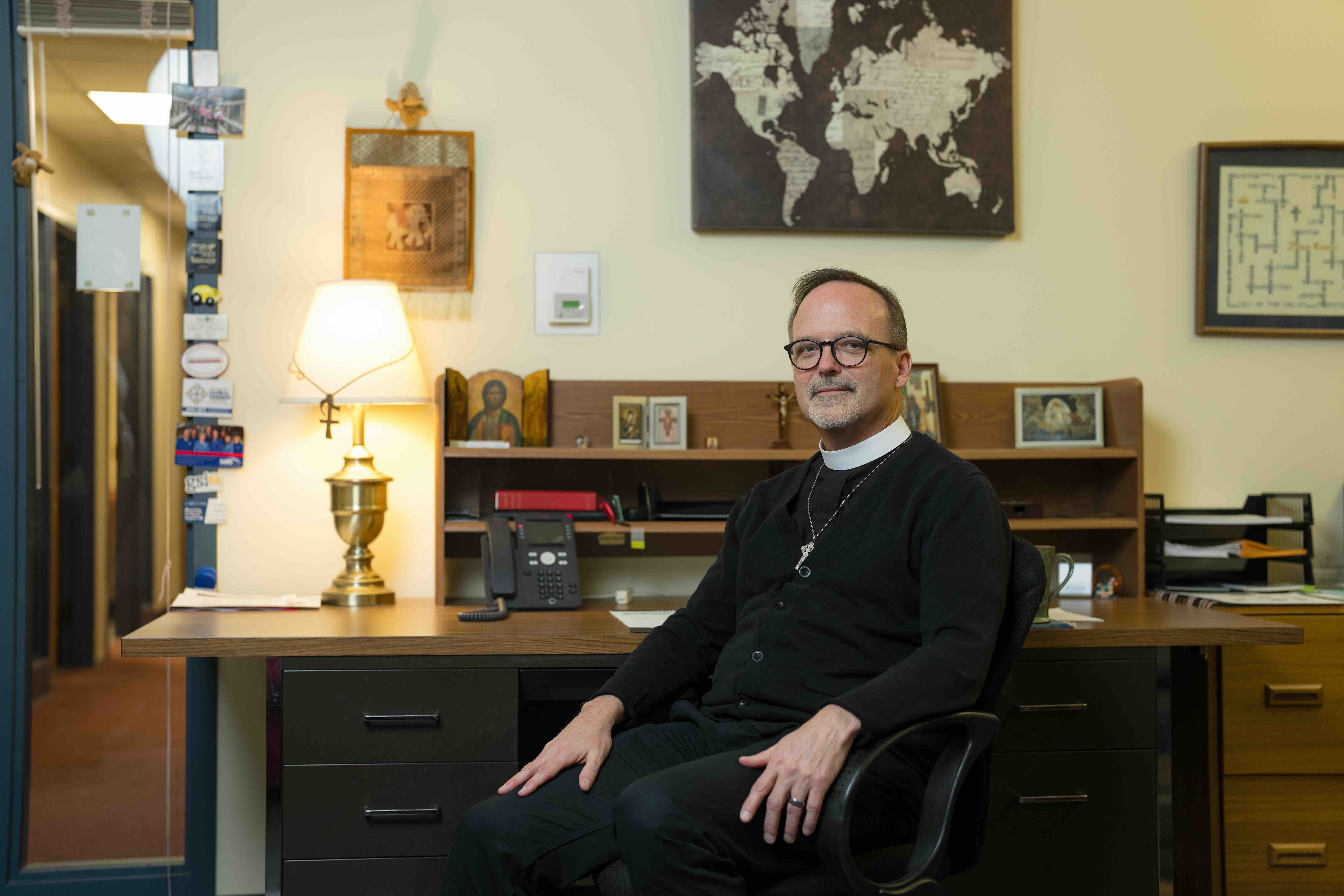 Man in clerical attire sits at a desk with a map on the wall and a lamp lit next to him. The room conveys a serene, professional atmosphere.