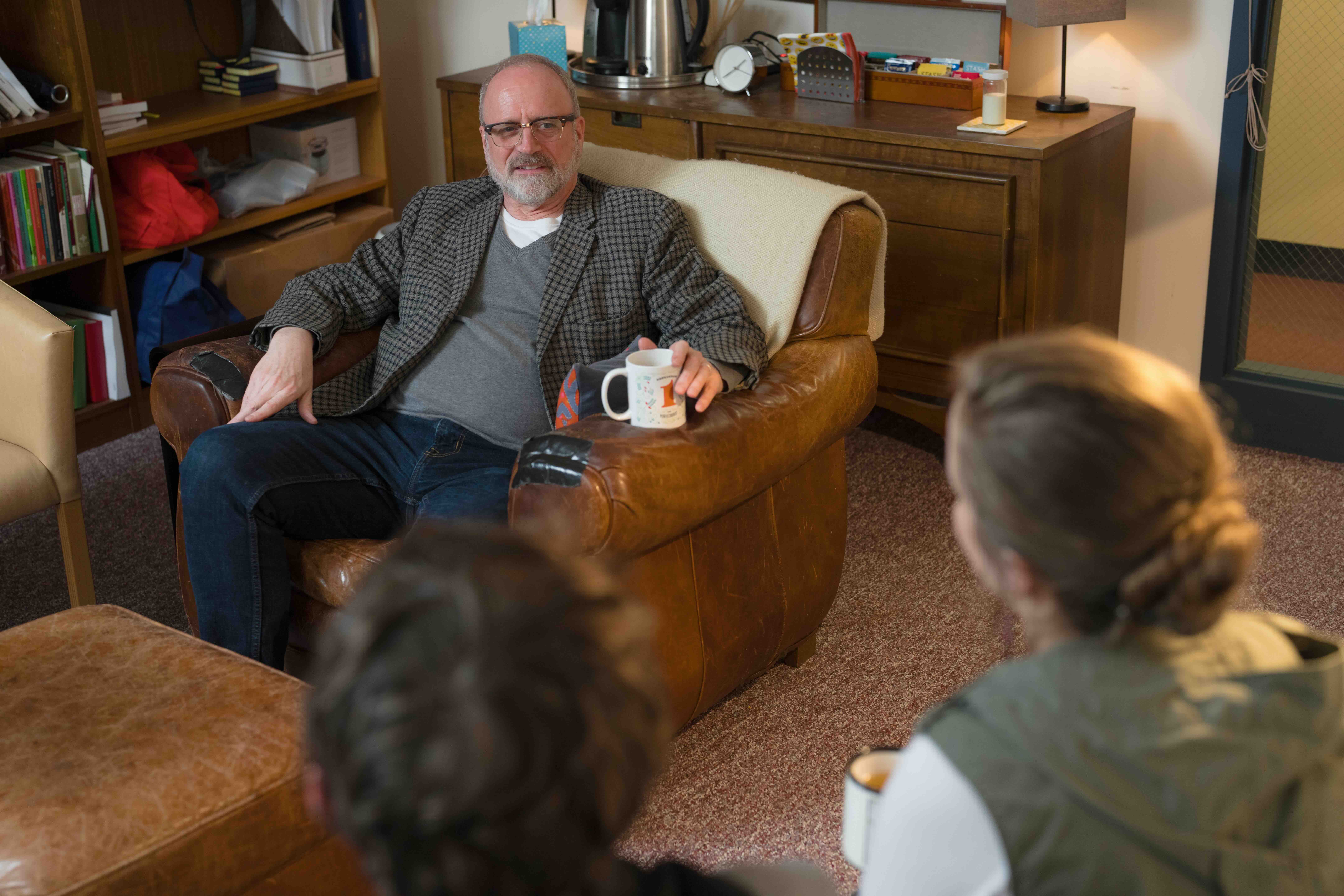 Middle-aged man with glasses sitting on a brown leather armchair holding a mug and talking to two people whose backs are to the camera in a cozy living room.
