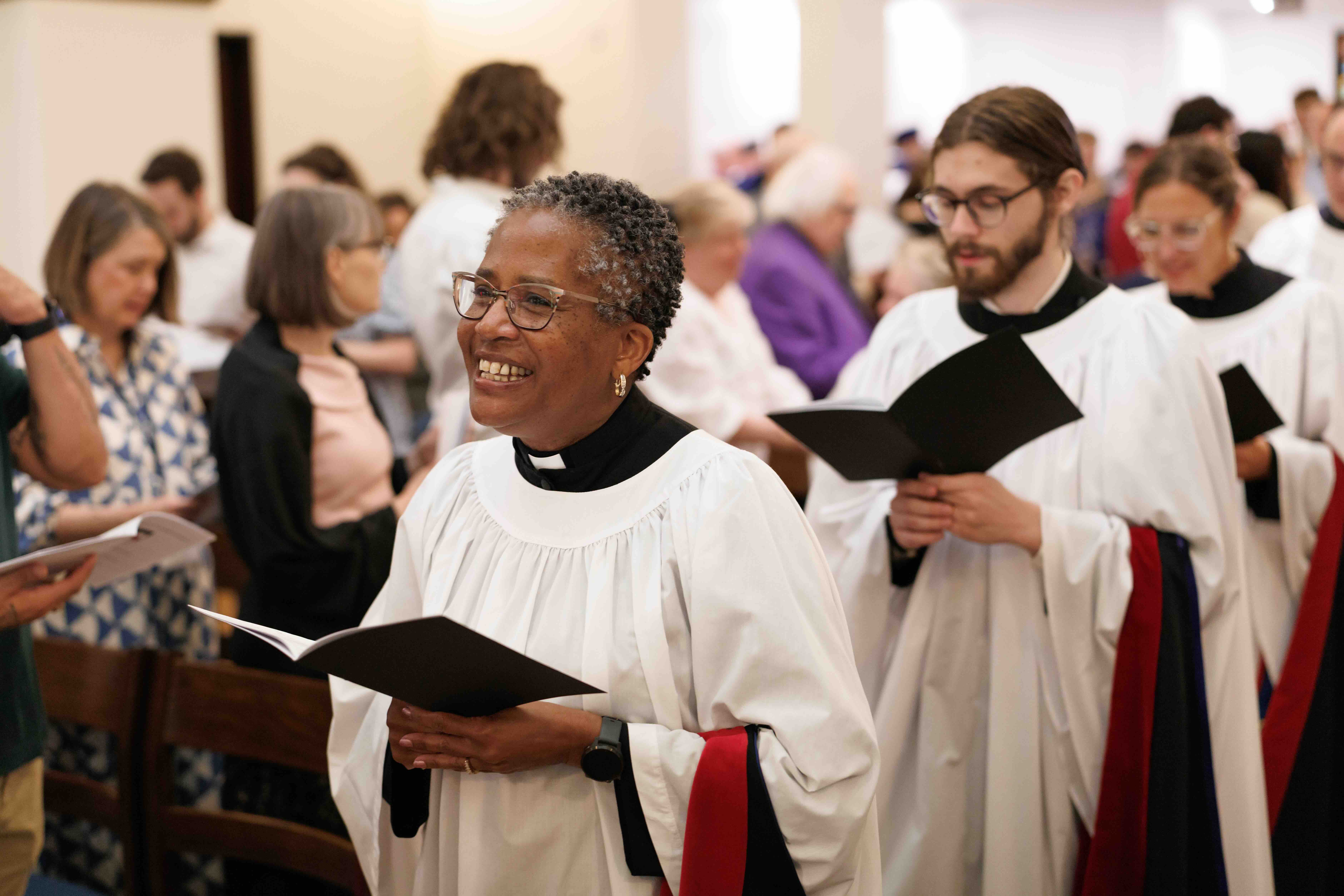 A joyful choir procession in a church setting. An individual in white vestments is smiling while singing from a booklet, surrounded by others in similar attire.